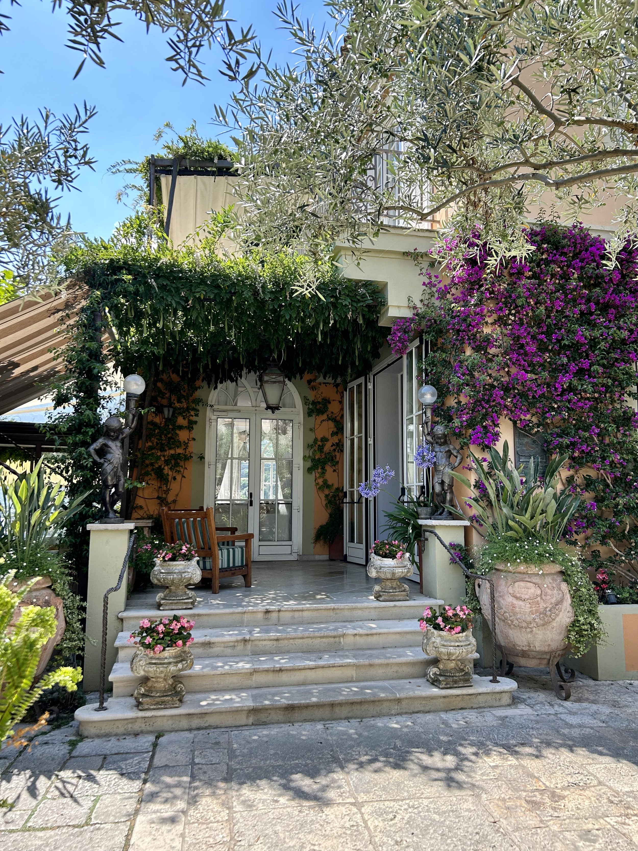 Entrance to a house with steps leading to a porch, decorated with potted flowers, statues, and lush greenery, including flowering vines and trees.