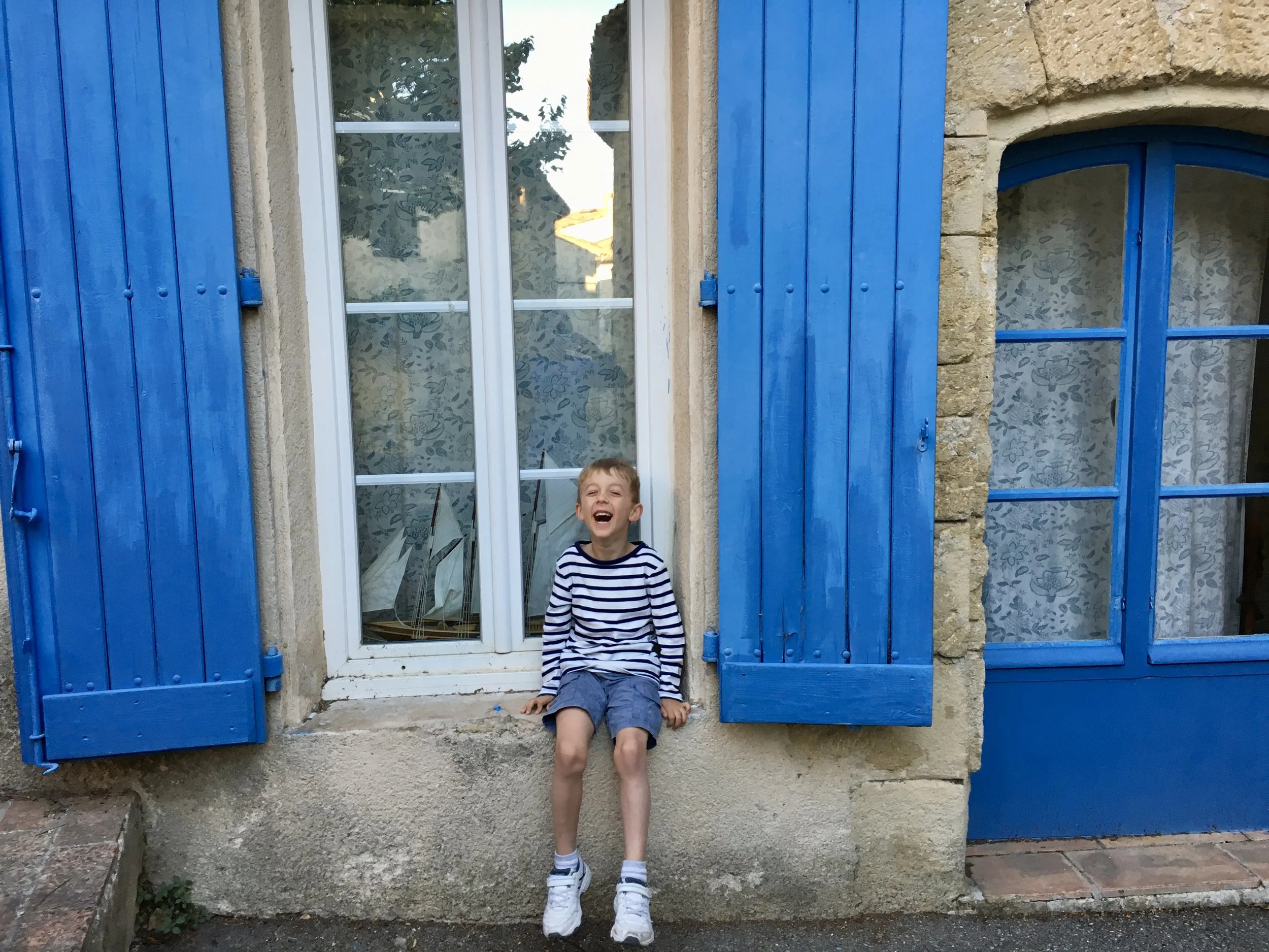A young boy with blonde hair, wearing a striped shirt, shorts, and sneakers, sitting on a windowsill outside a building with bright blue shutters and stone walls, laughing.