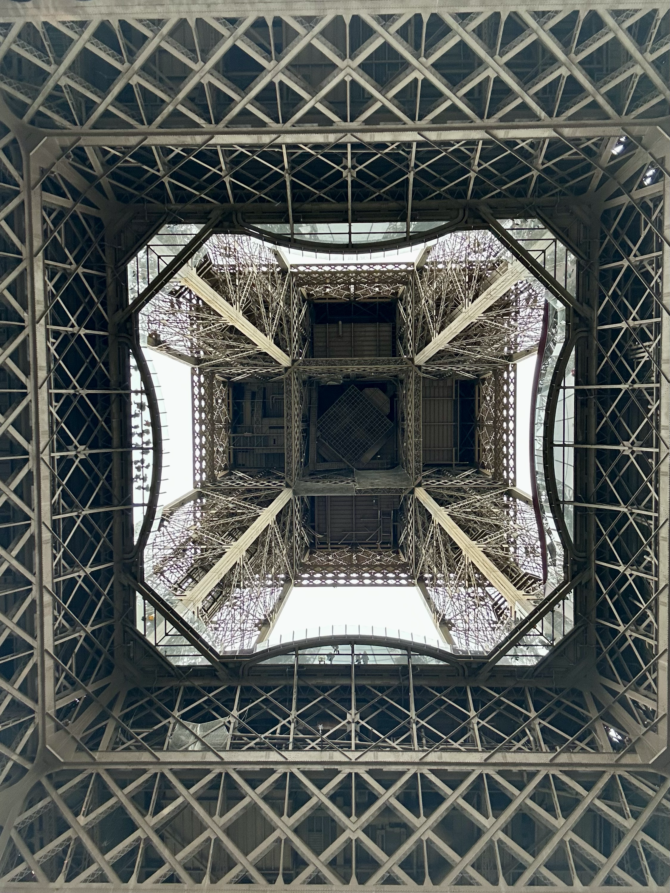 Looking up at the Eiffel Tower from directly underneath, showcasing the intricate lattice ironwork and square-shaped structure with a view of the sky through the openings.