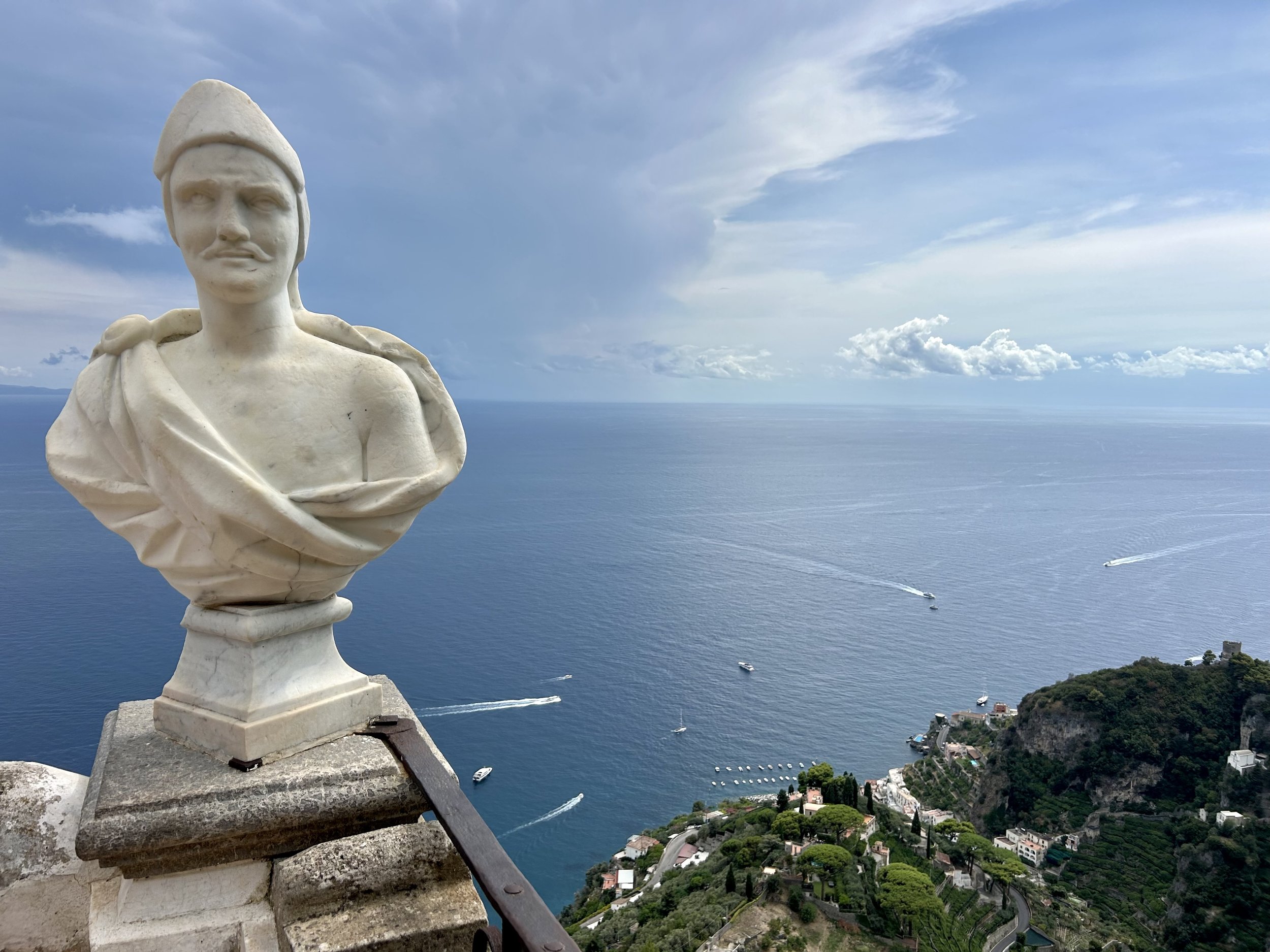 A marble bust of a man wearing a hat, positioned on a ledge overlooking a coastal landscape with boats on the water and a hillside town below.