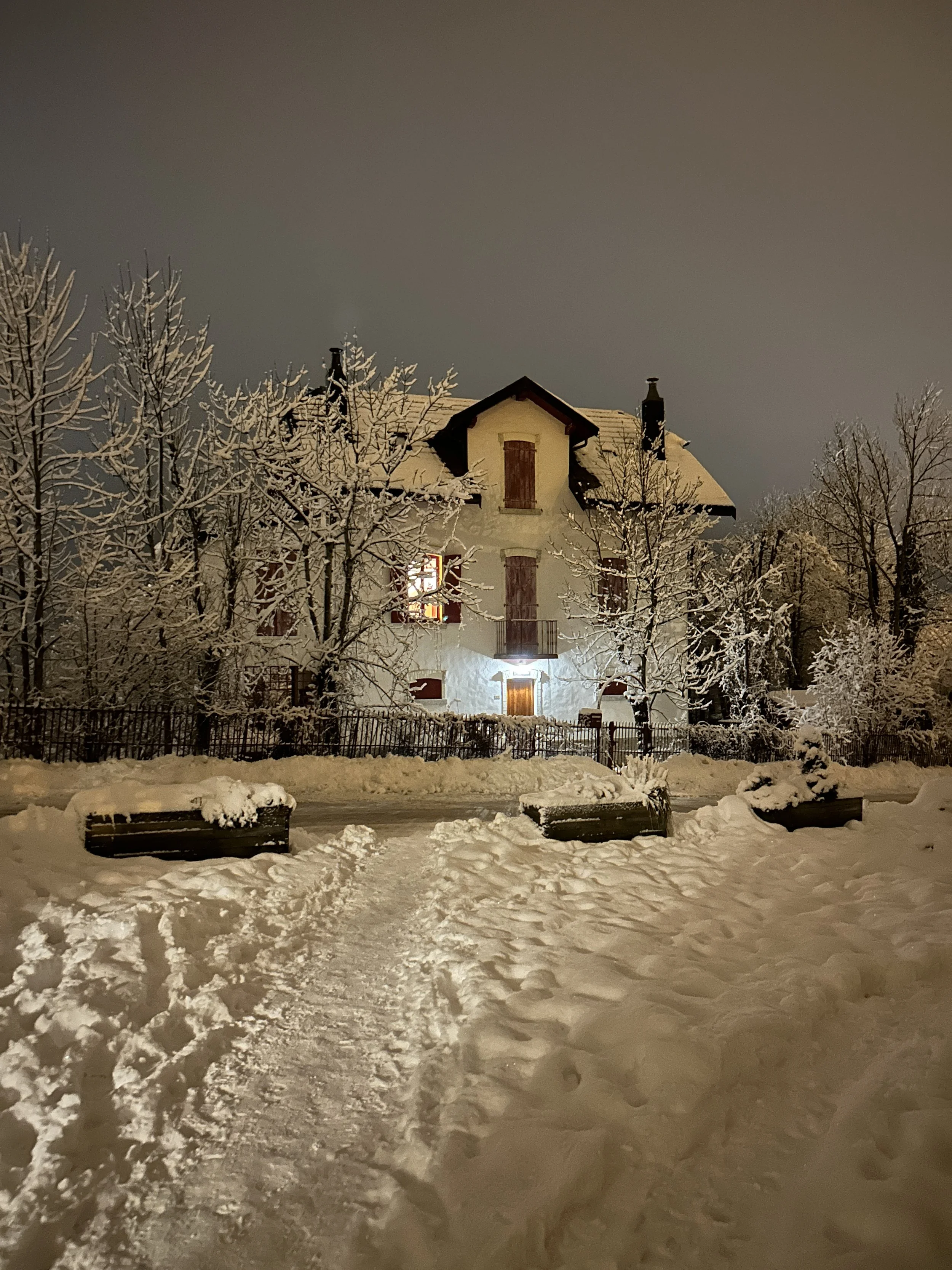 Snow-covered house at night with illuminated windows, surrounded by snow-laden trees and benches, footprints in the snow leading to the house.