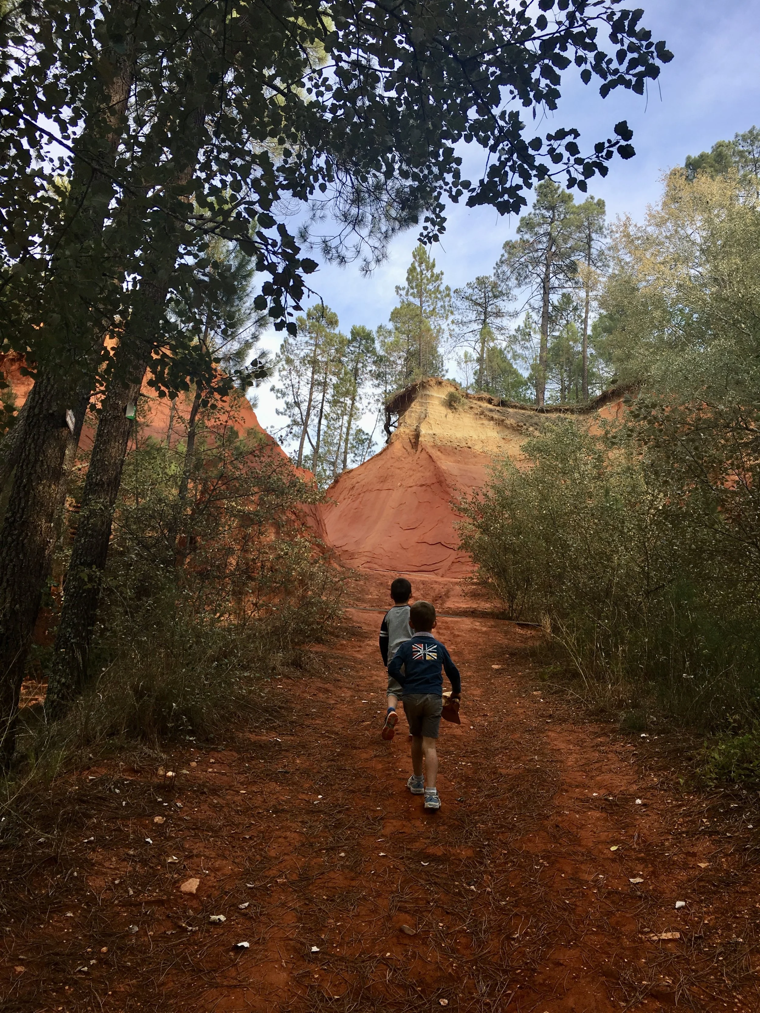 Two kids hiking on a red dirt trail surrounded by trees, with a sandstone formation in the background and blue sky above.