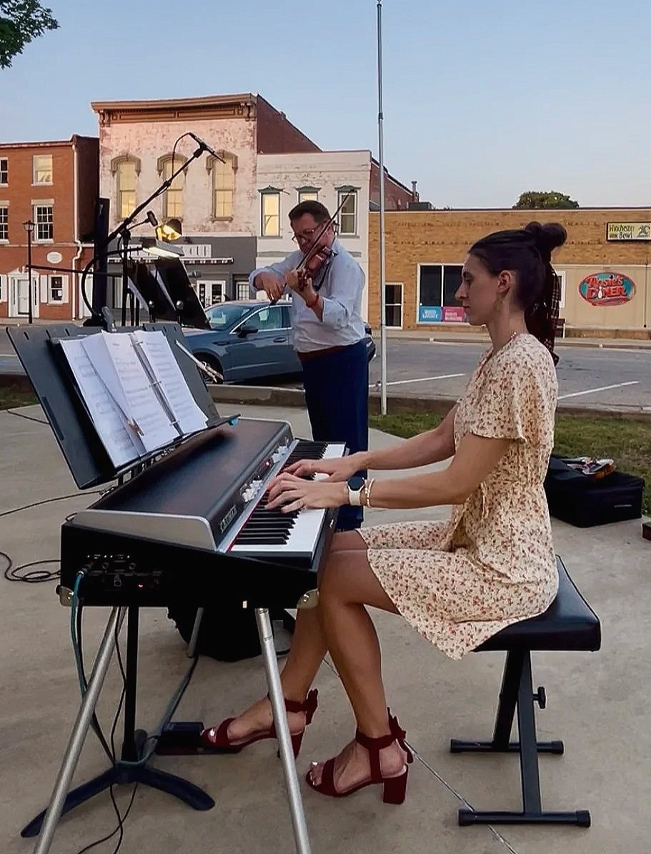 A woman in a floral dress playing a keyboard while a man behind her on the street plays a violin. They are outside with buildings and parked cars in the background.