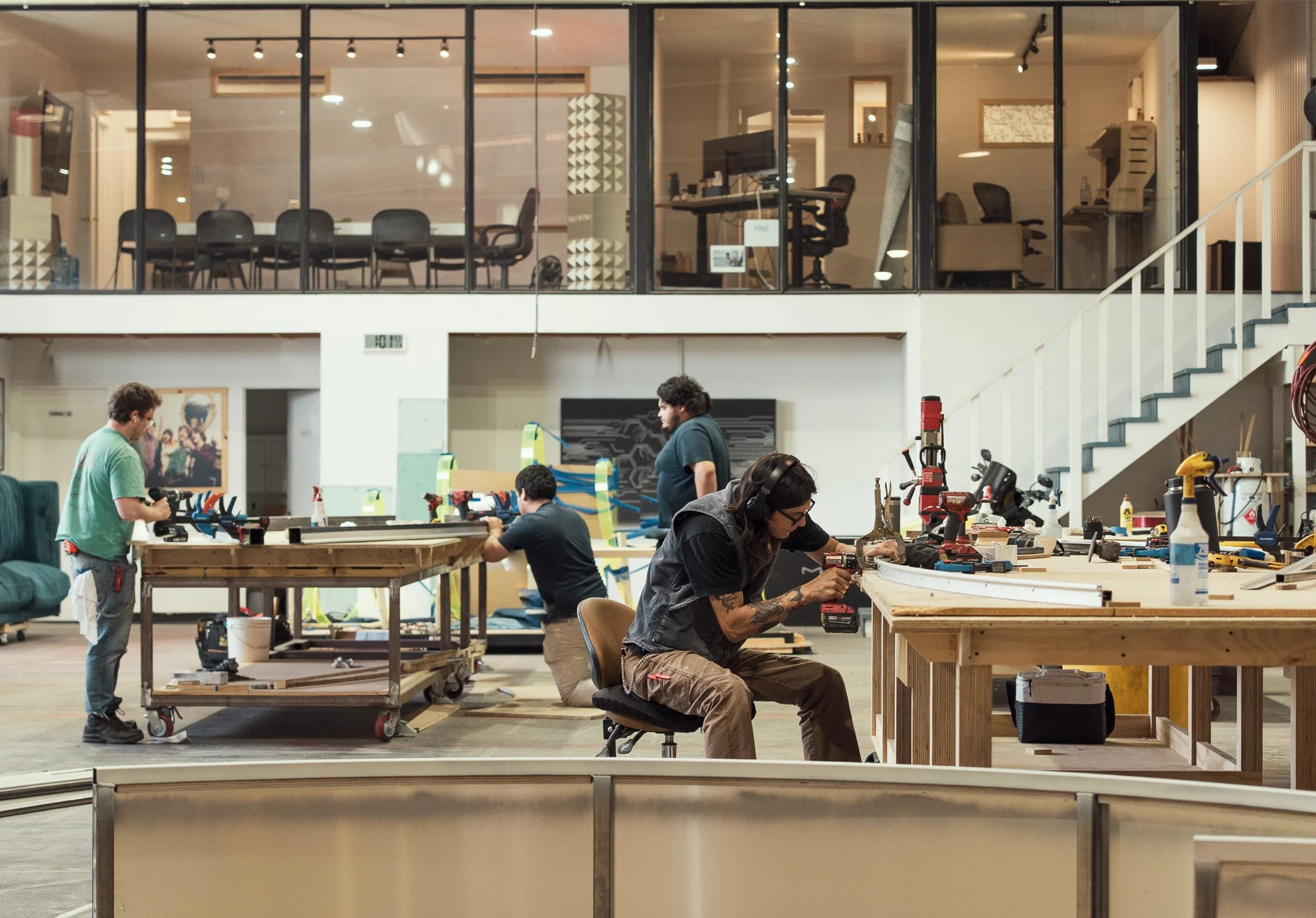 Experienced and professional expert builders working on big fabrication projects for events and activations in a clean workshop with a large, multi-level interior and upstairs offices, conference room, and design studio visible through glass walls.