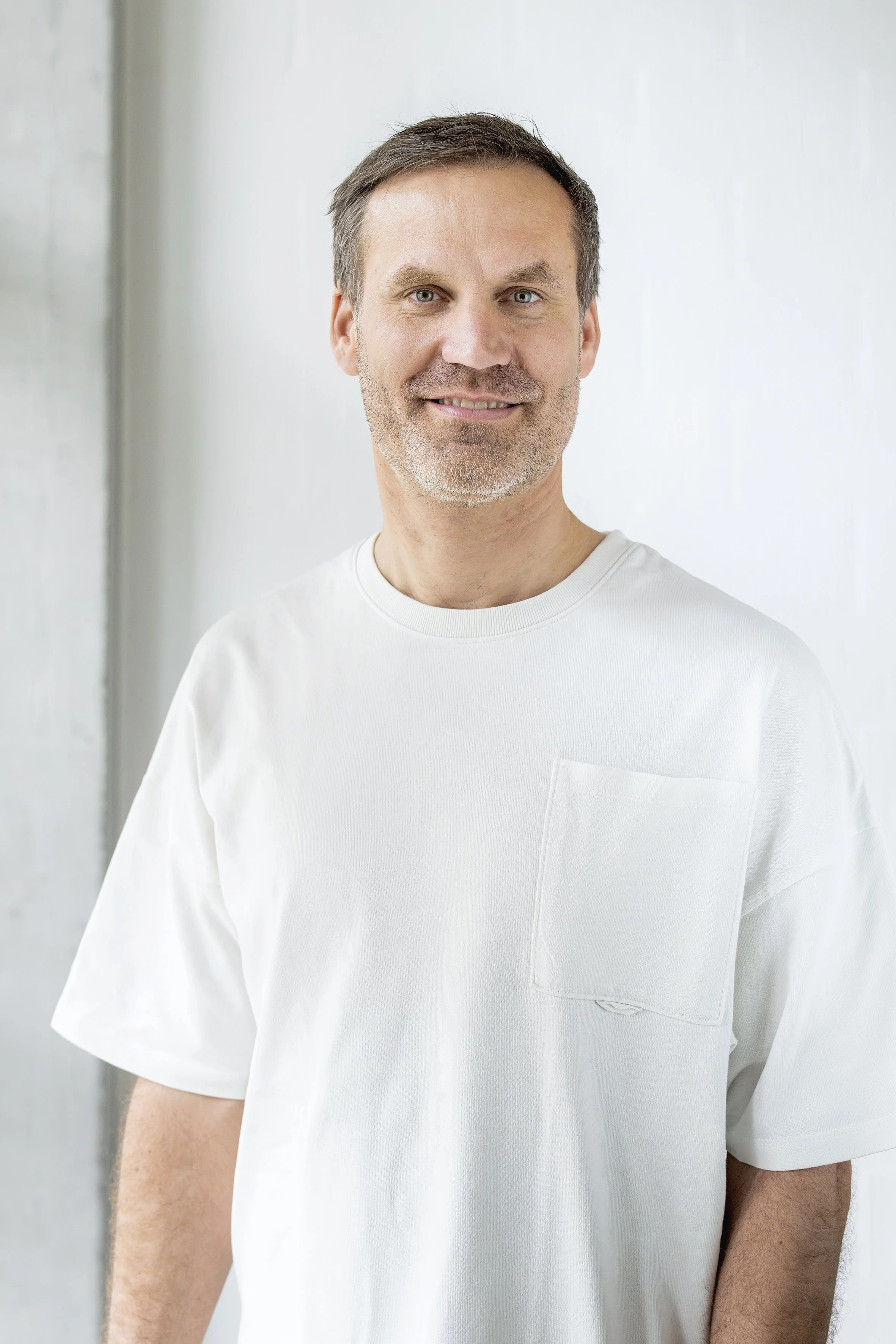 Ein Mann mittleren Alters mit kurzen braunen Haaren, grauem Bart und blauen Augen lächelt, trägt ein schlichtes weißes T-Shirt und steht in einem Haus vor einem schlichten hellen Hintergrund.