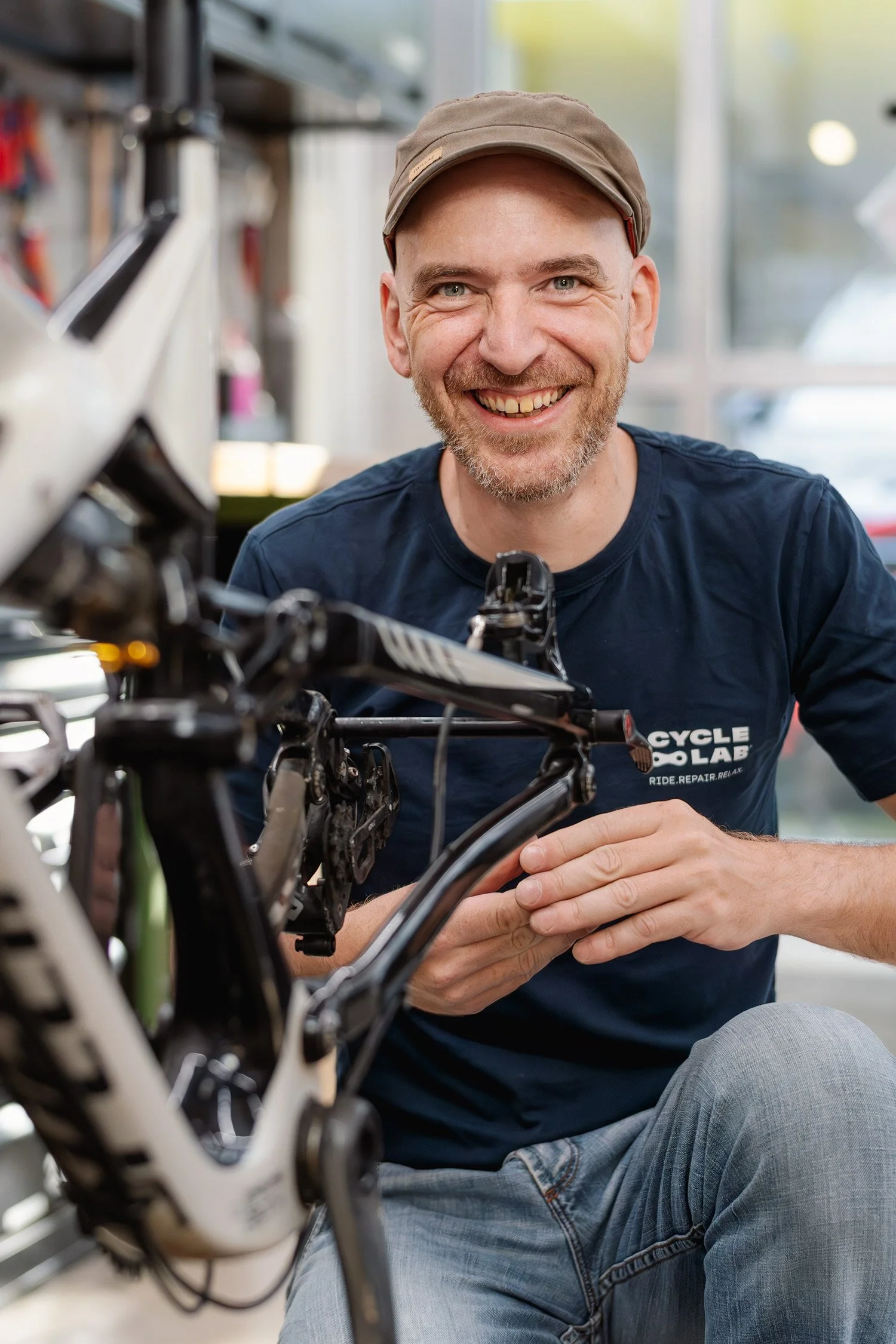 Ein lächelnder Mann, der in einer Werkstatt an einem Fahrrad arbeitet, trägt eine Mütze und ein marineblaues T-Shirt.
