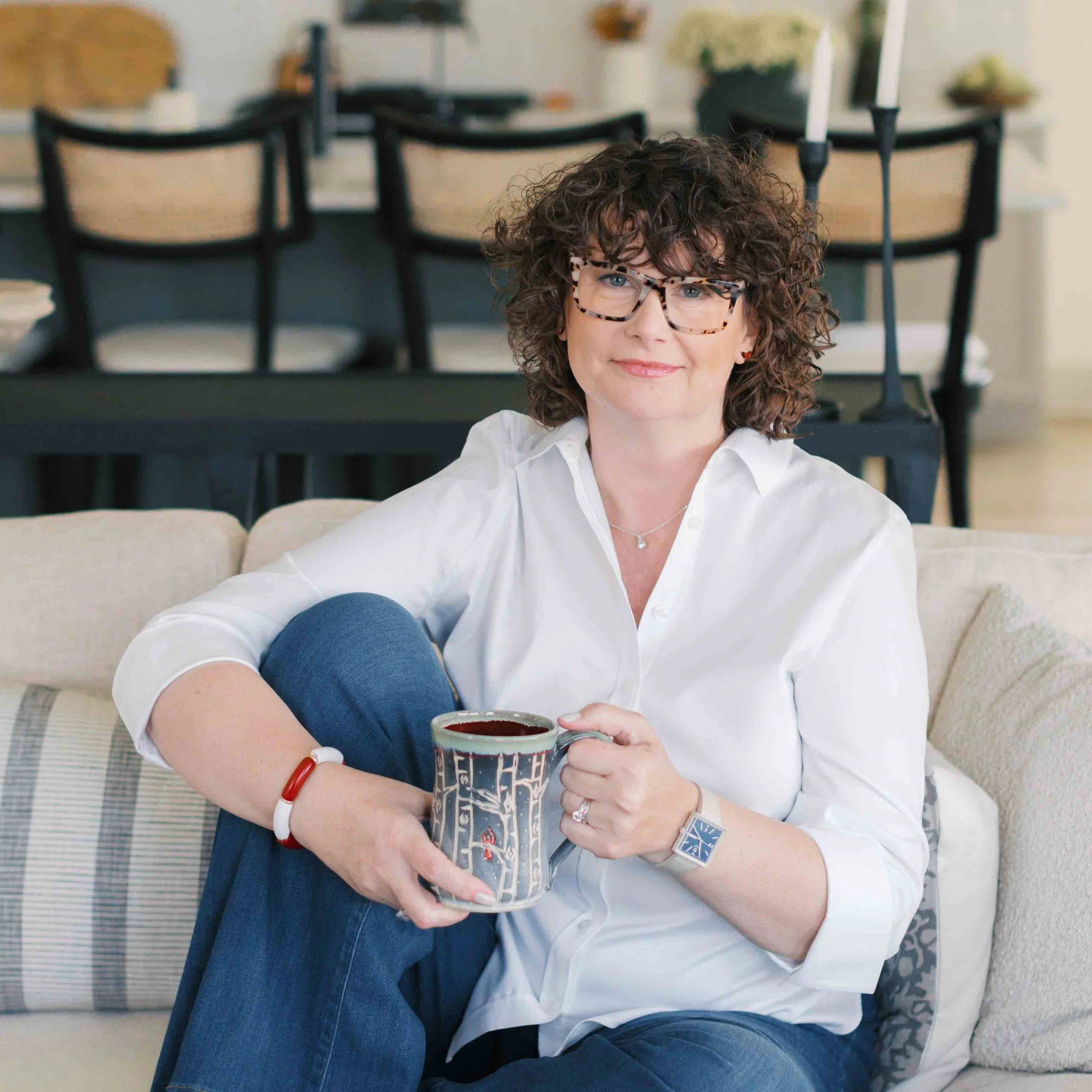 Nichole, a woman with curly hair and glasses sitting on a beige couch, holding a patterned coffee mug filled with coffee, wearing a white blouse and blue jeans, in a cozy living room.