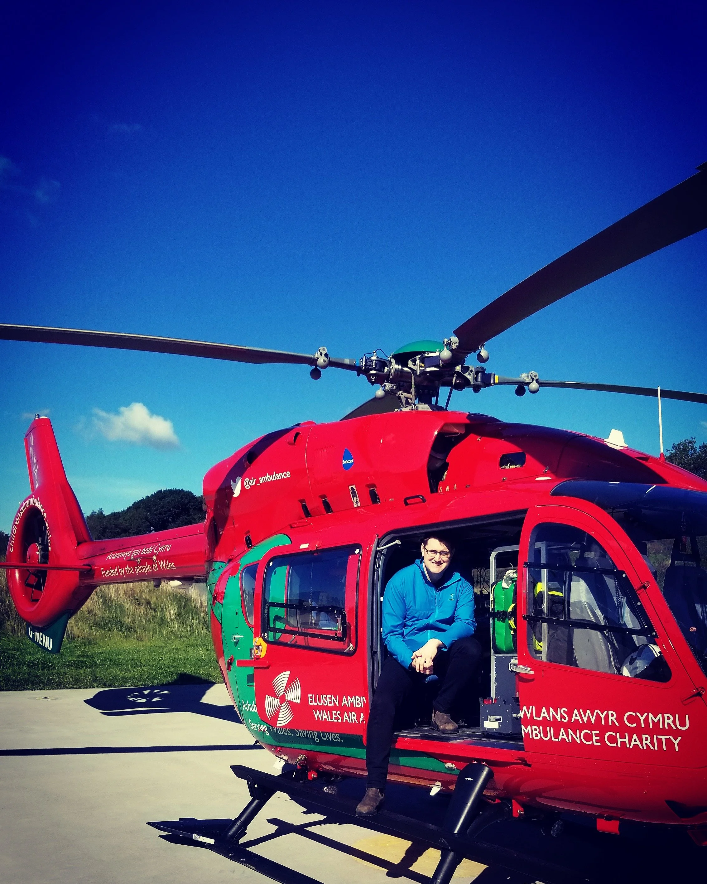 A red helicopter with a person sitting in the open door, smiling. The helicopter has logos and writing indicating it's an ambulance for Wales Air Ambulance.