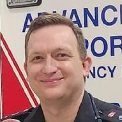 A man smiling in front of a sign with the words 'Advance' and 'Emergency' visible.