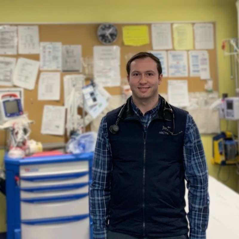 A male doctor or healthcare professional inside a medical clinic or hospital, standing in front of a bulletin board with various papers and notices, wearing a stethoscope around his neck, a black vest, and a blue plaid shirt.