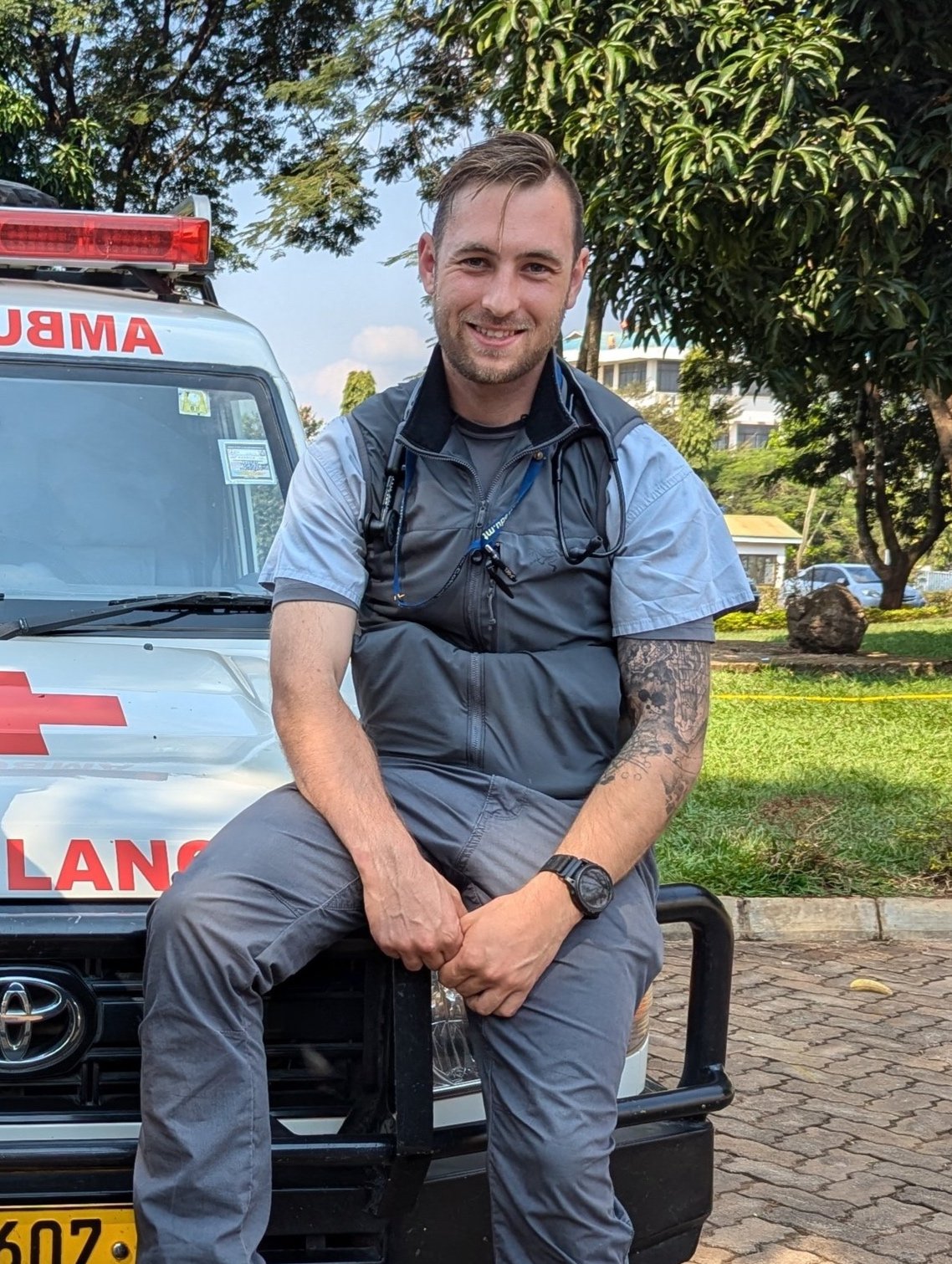 A smiling young man in paramedic uniform sitting on the front bumper of an ambulance with trees and buildings in the background.