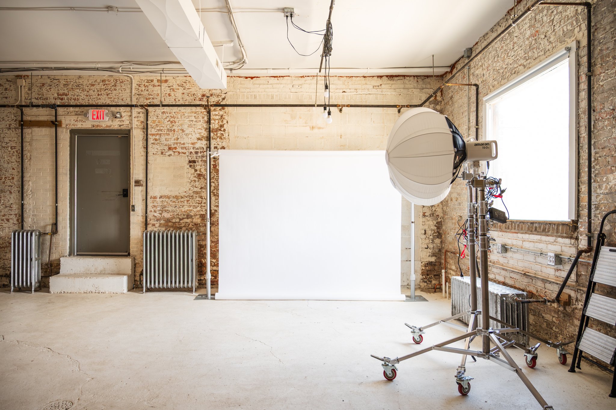 AH AH's industrial-style photo and film studio with exposed brick walls and large windows, equipment including a large softbox on a rolling stand, and a backdrop stand with a white backdrop.