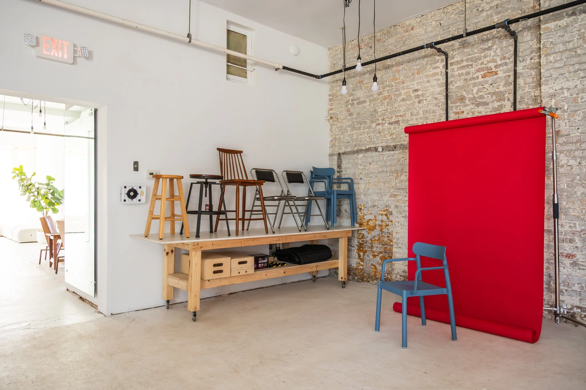 Interior of a AH AH's photography studio with a white brick wall and a red backdrop roll next to a row of prop chairs.