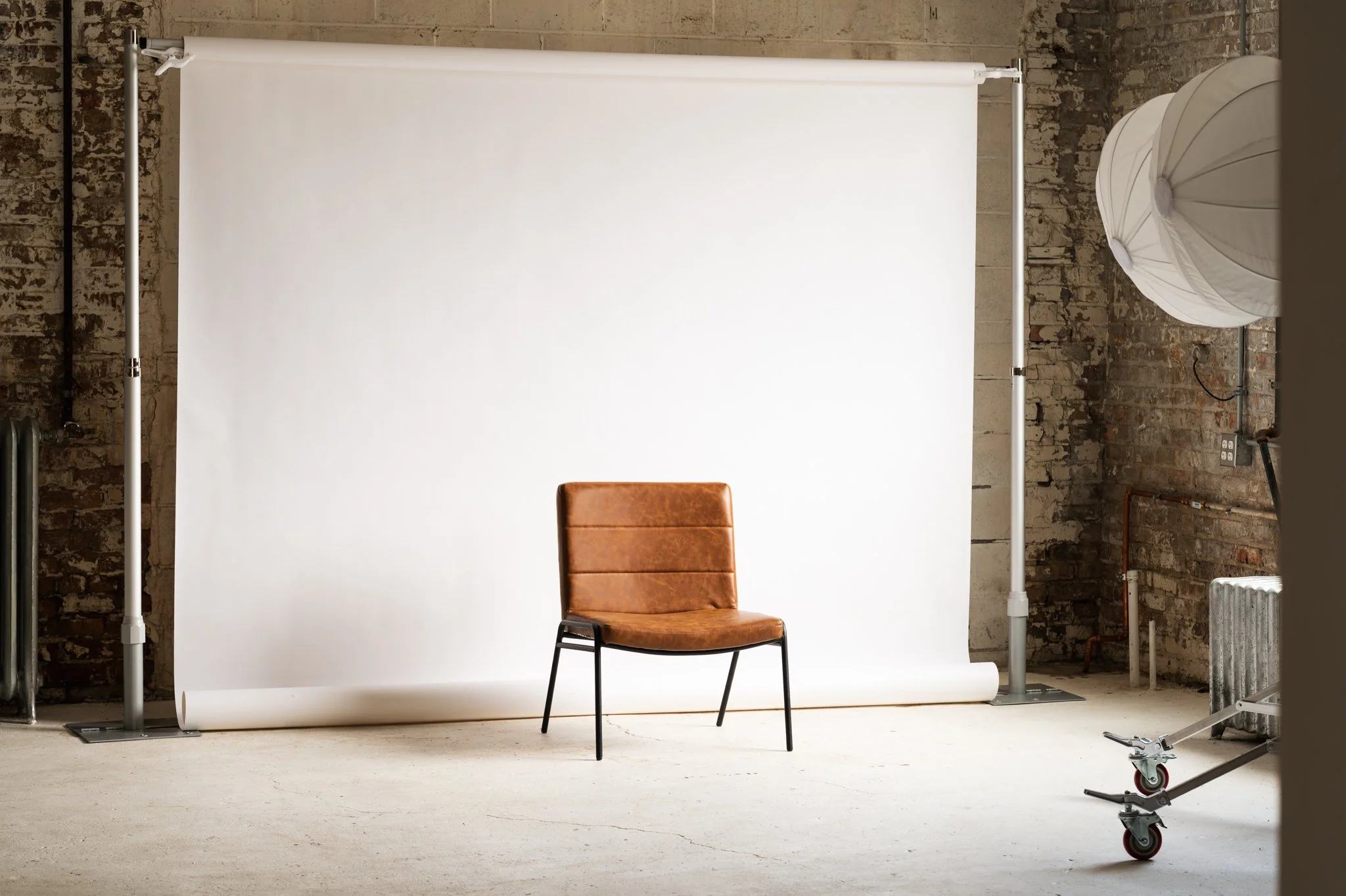 AH AH's photography studio setup featuring a large white backdrop on a stand, a vintage brown leather chair, and professional studio lighting equipment in a room with exposed brick walls.