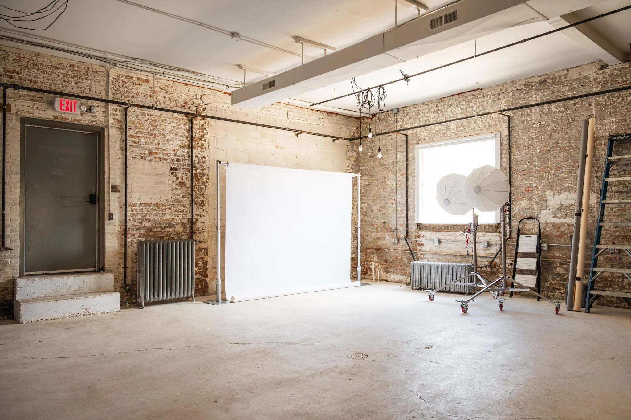 AH AH's industrial-style photo and film studio with exposed brick walls and large windows, equipment including a large softbox on a rolling stand, and a backdrop stand with a white backdrop.