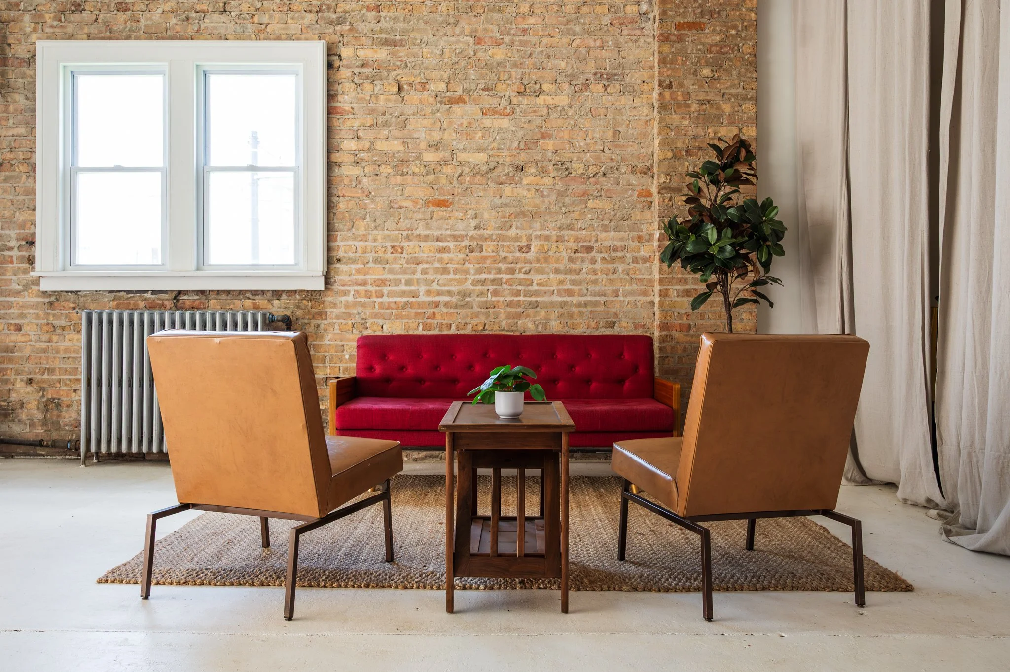 A sitting area set within AH AH's photo and film studio featuring an exposed brick wall, a red cushioned sofa, two tan leather armchairs, and a wooden table. Overhead, there are exposed light bulbs hanging from black cords.