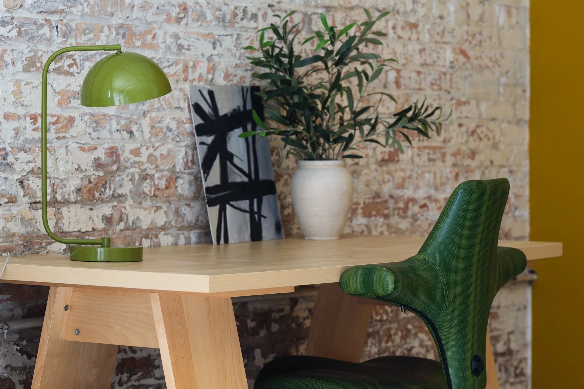 A workspace setup in the back area of AH AH's photography studio featuring a wooden desk with a green desk lamp, a potted plant, a black and white abstract painting, and a green chair in front of an exposed brick wall.