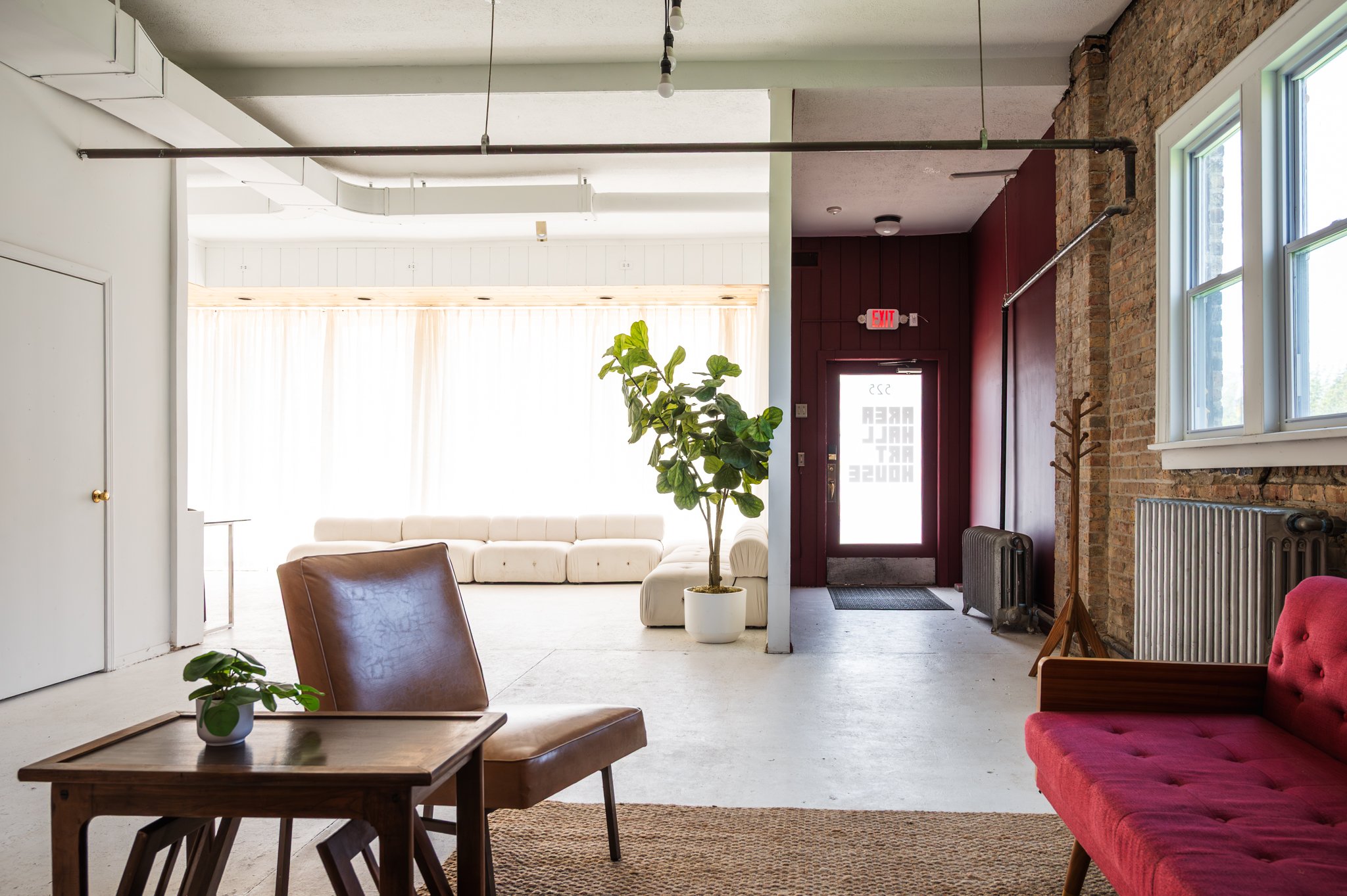 A sitting area set within AH AH's photo and film studio featuring an exposed brick wall, a red cushioned sofa, two tan leather armchairs, and a wooden table. Overhead, there are exposed light bulbs hanging from black cords.