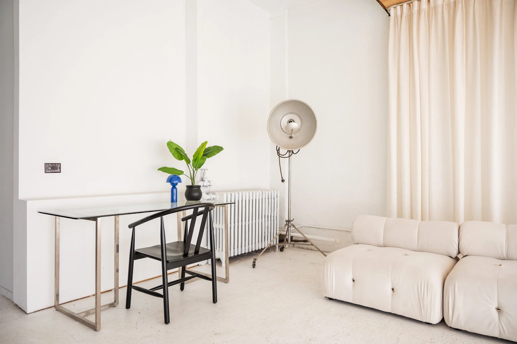 The front area of Area Hall Art House that displays a simple workspace setup with a black modern chair, a glass desk with an accent light and plant.