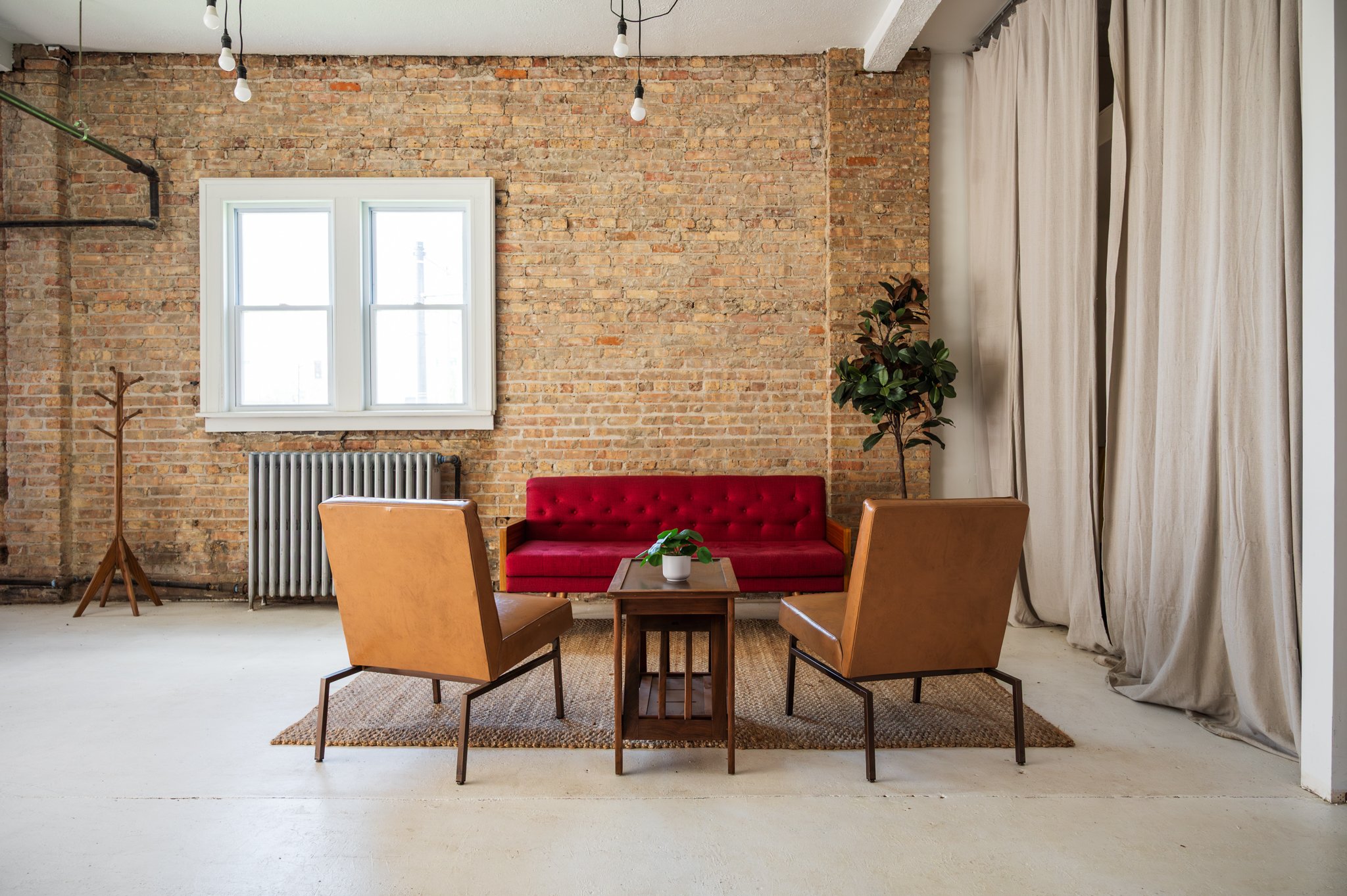 A sitting area set within AH AH's photo and film studio featuring an exposed brick wall, a red cushioned sofa, two tan leather armchairs, and a wooden table. Overhead, there are exposed light bulbs hanging from black cords.