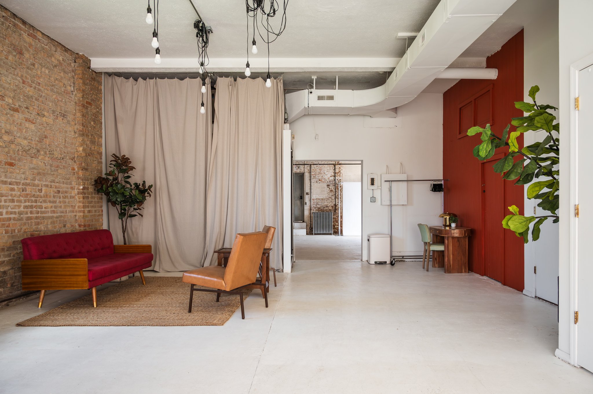 A sitting area set within AH AH's photo and film studio featuring a red cushioned sofa, two tan leather armchairs, and a wooden table. Overhead, there are exposed light bulbs hanging from black cords.