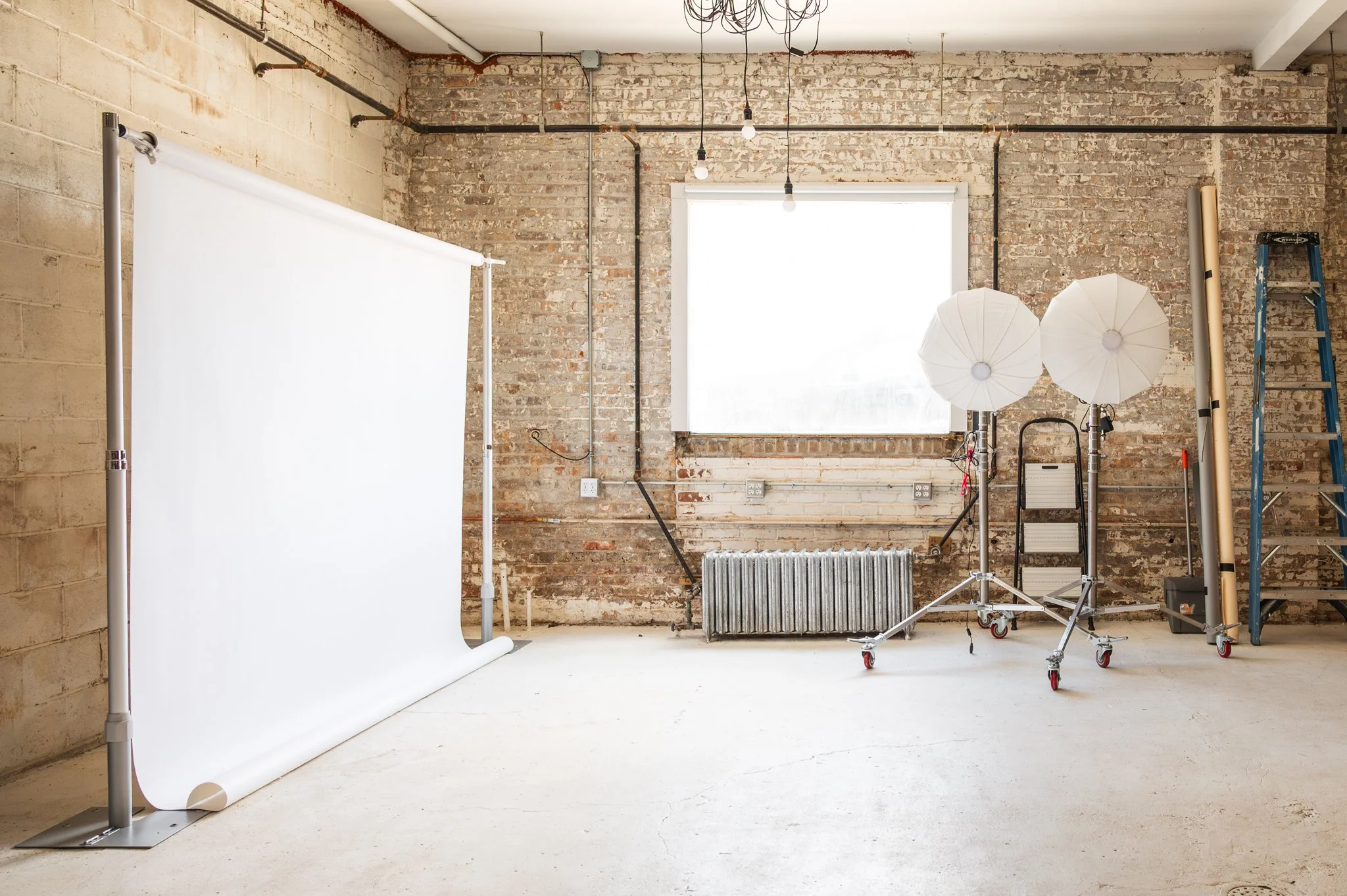 AH AH's back photography studio space with a white backdrop on a stand, studio lights with softboxes, various photo gear and equipment that is including in each booking.