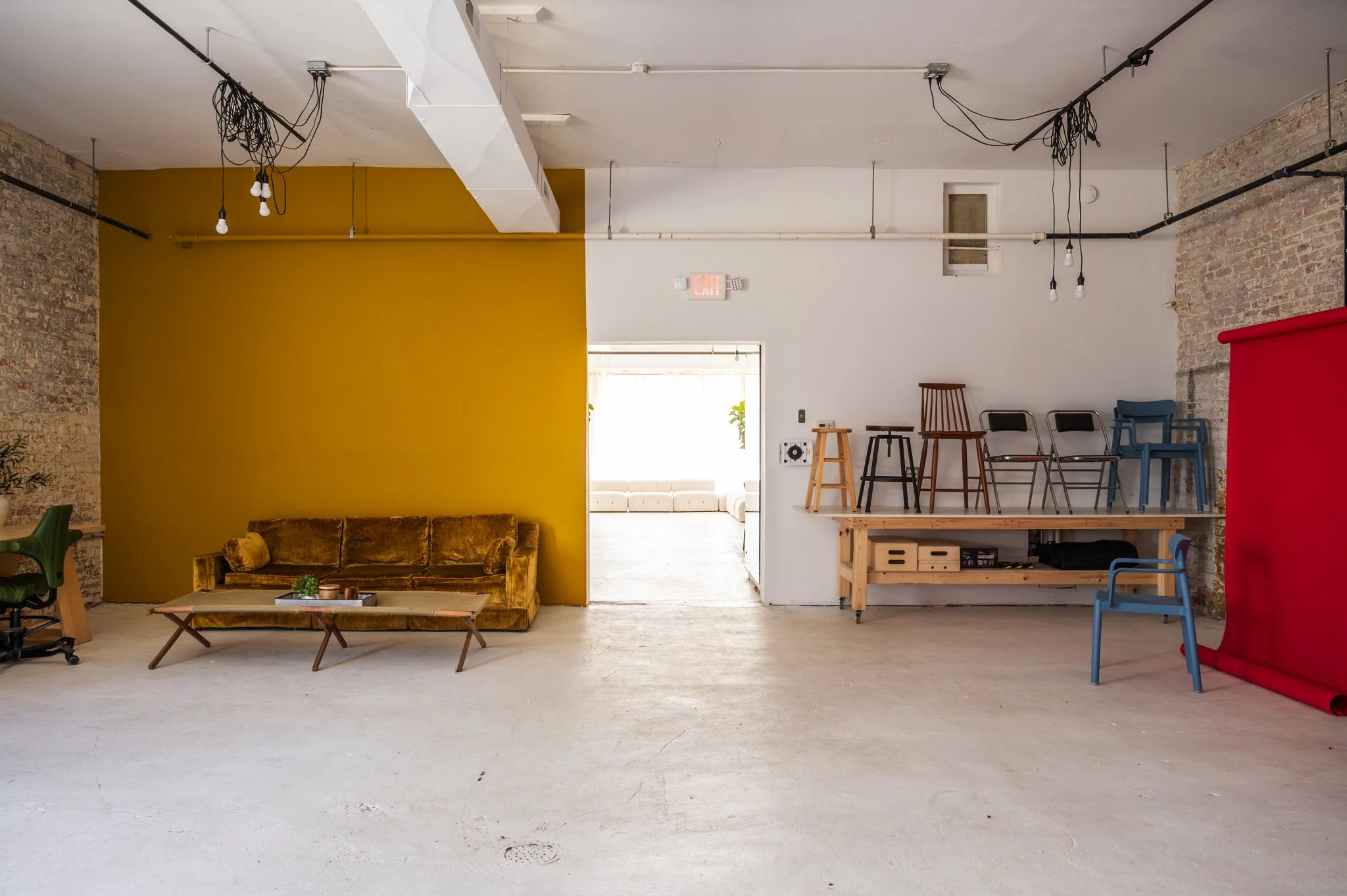 Modern, industrial-style interior of Area Hall Art House, a photography and film studio with exposed brick walls, a mustard yellow accent wall, and a seating area with a vintage velvet sofa and coffee table, and a red backdrop on the right side.