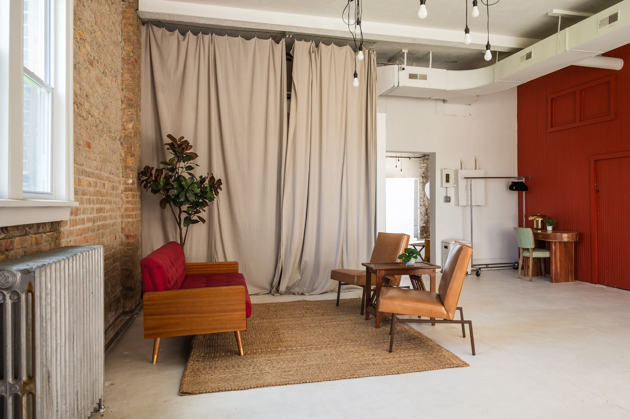 A sitting area set within AH AH's photo and film studio featuring a red cushioned sofa, two tan leather armchairs, and a wooden table. Overhead, there are exposed light bulbs hanging from black cords.