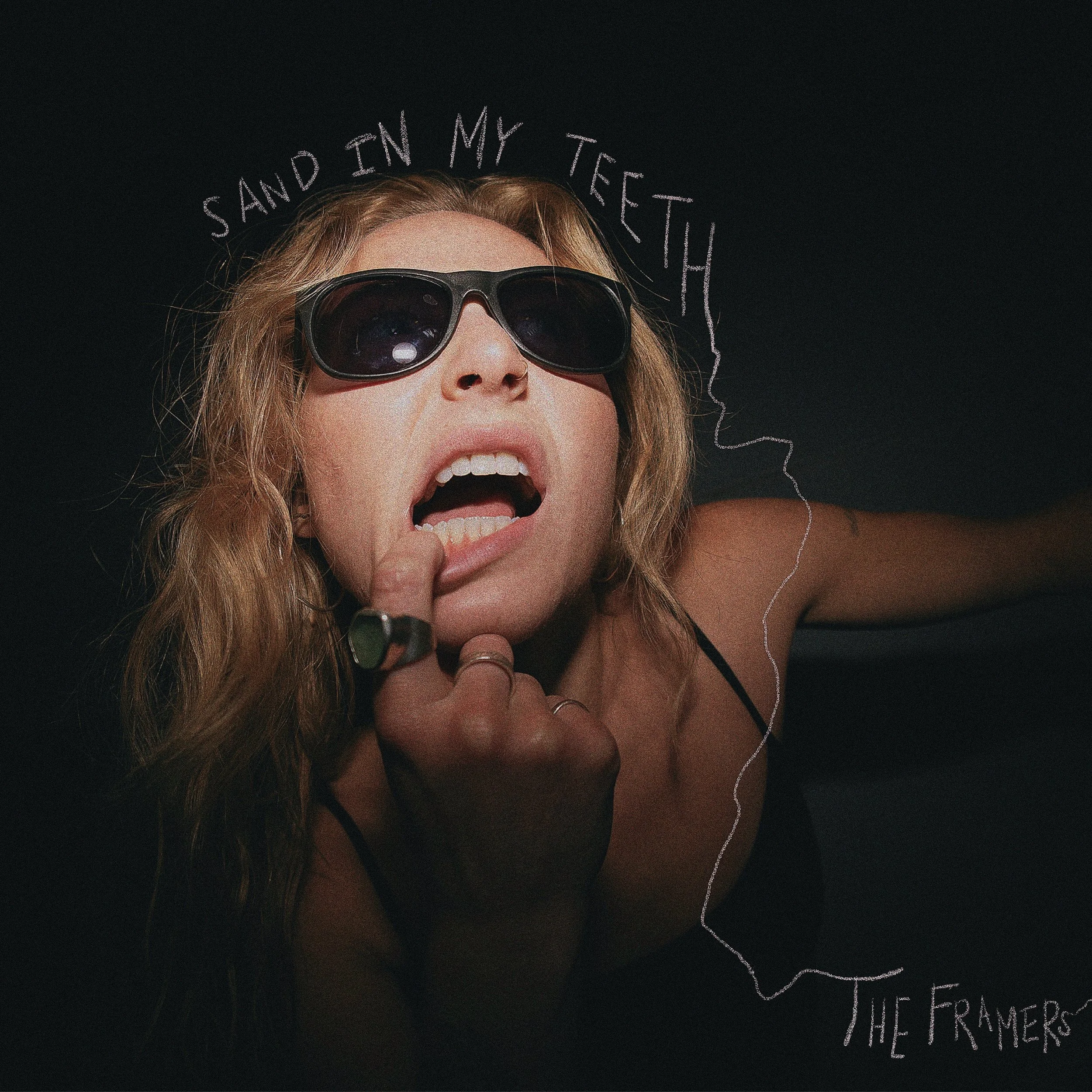 A woman with wavy hair wearing sunglasses is holding her finger to her mouth. The text 'Sand in my teeth' is written above her head in a doodle style. This is the cover art for a song named Sand in My Teeth, written and performed by The Framers.