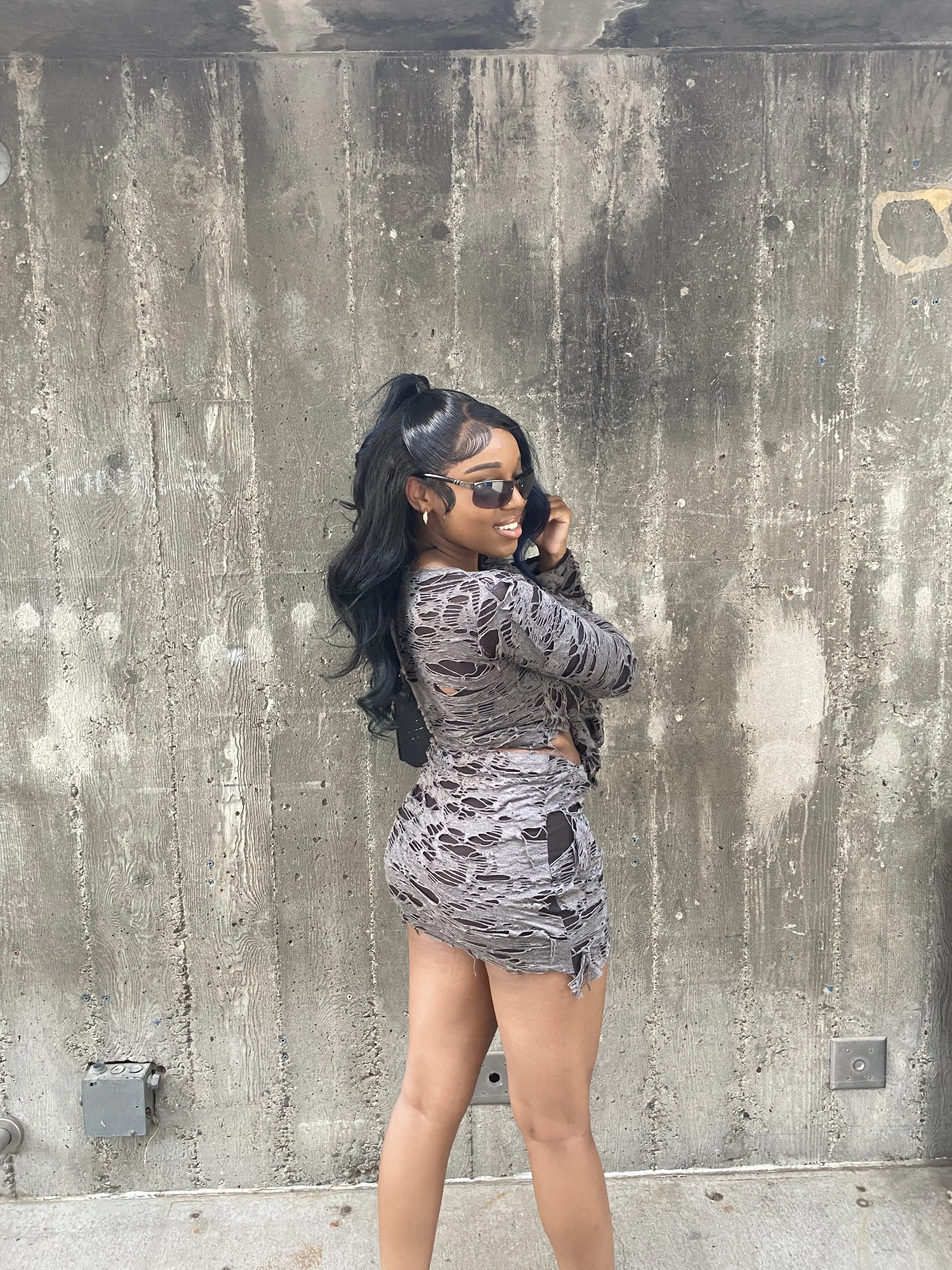 A young woman with long black hair, wearing sunglasses and a torn, fitted dress, stands against a concrete wall, smiling and posing with a hand near her face.
