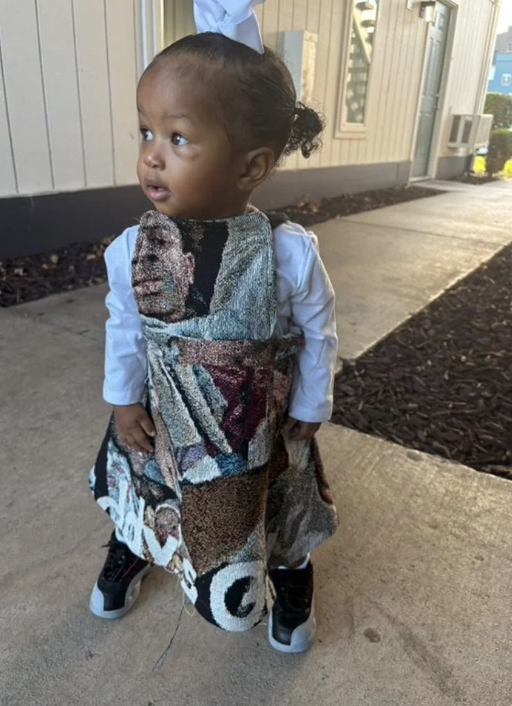 Young child standing outdoors, wearing a large tapestry-like bag over their shoulder, a white shirt, and black sneakers, with a building and sidewalk in the background.