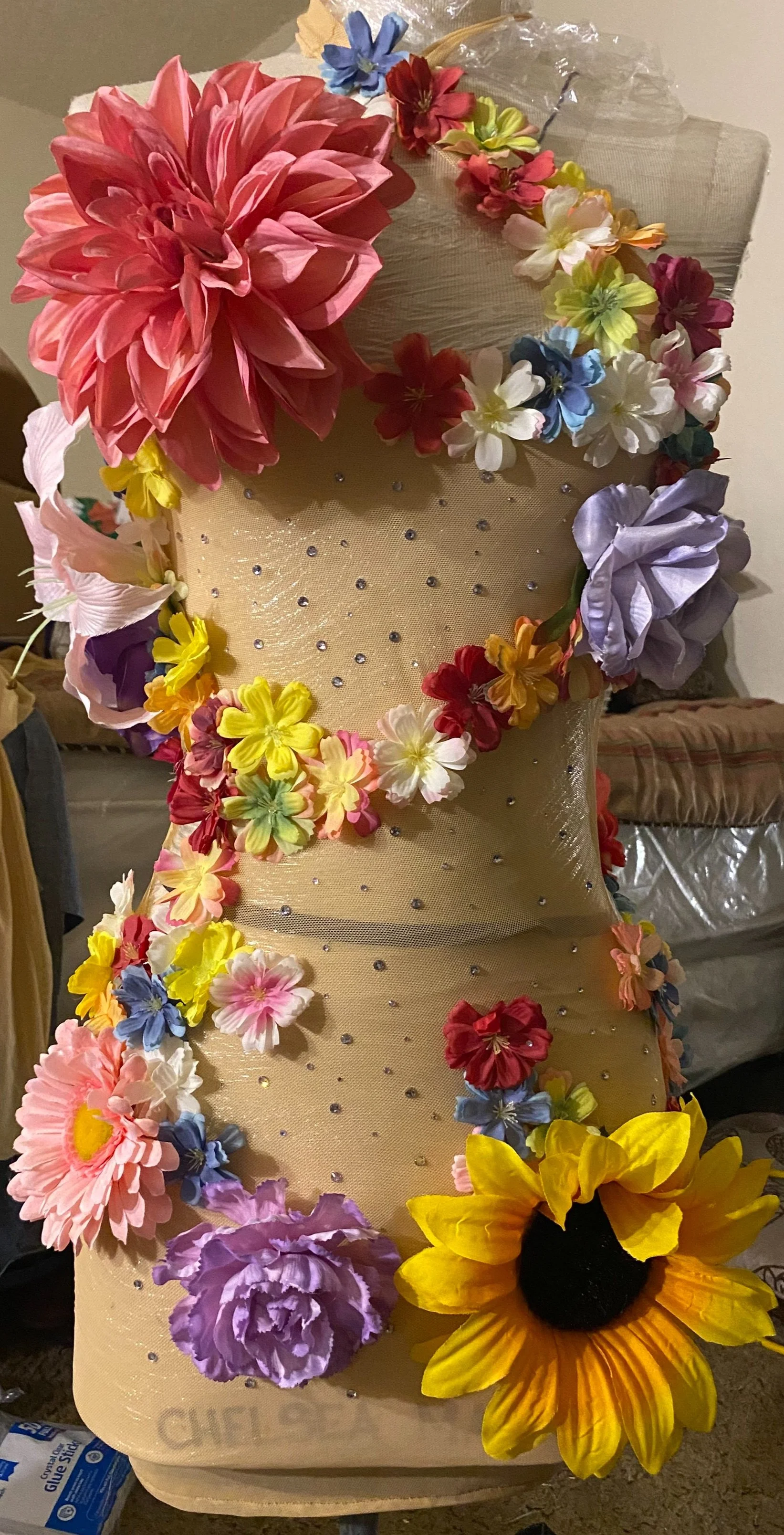 Decorative mannequin bust covered in beige fabric, adorned with colorful artificial flowers, including sunflowers, roses, daisies, and other blooms, with small rhinestones scattered across the fabric.