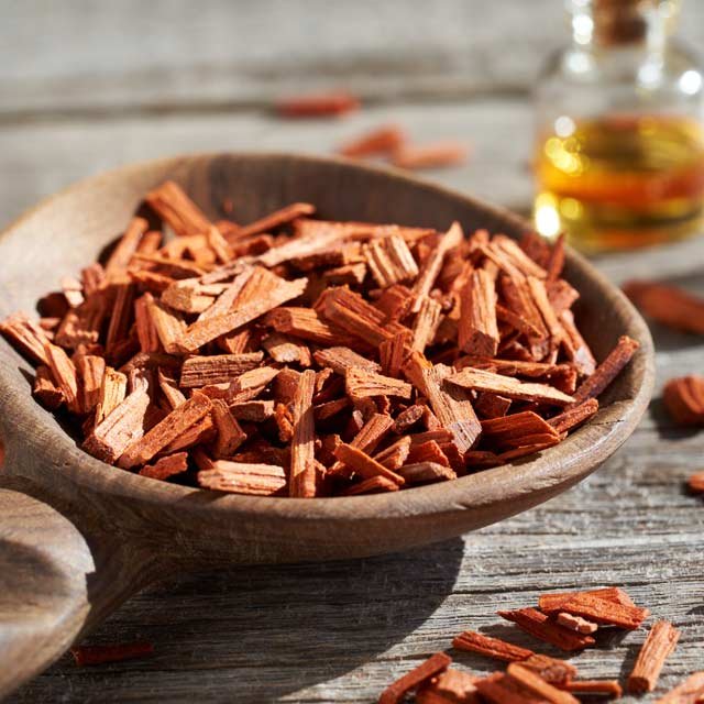 A wooden bowl filled with chopped cedar bark on a rustic wooden surface, with some pieces scattered around, and a glass bottle of oil in the background.