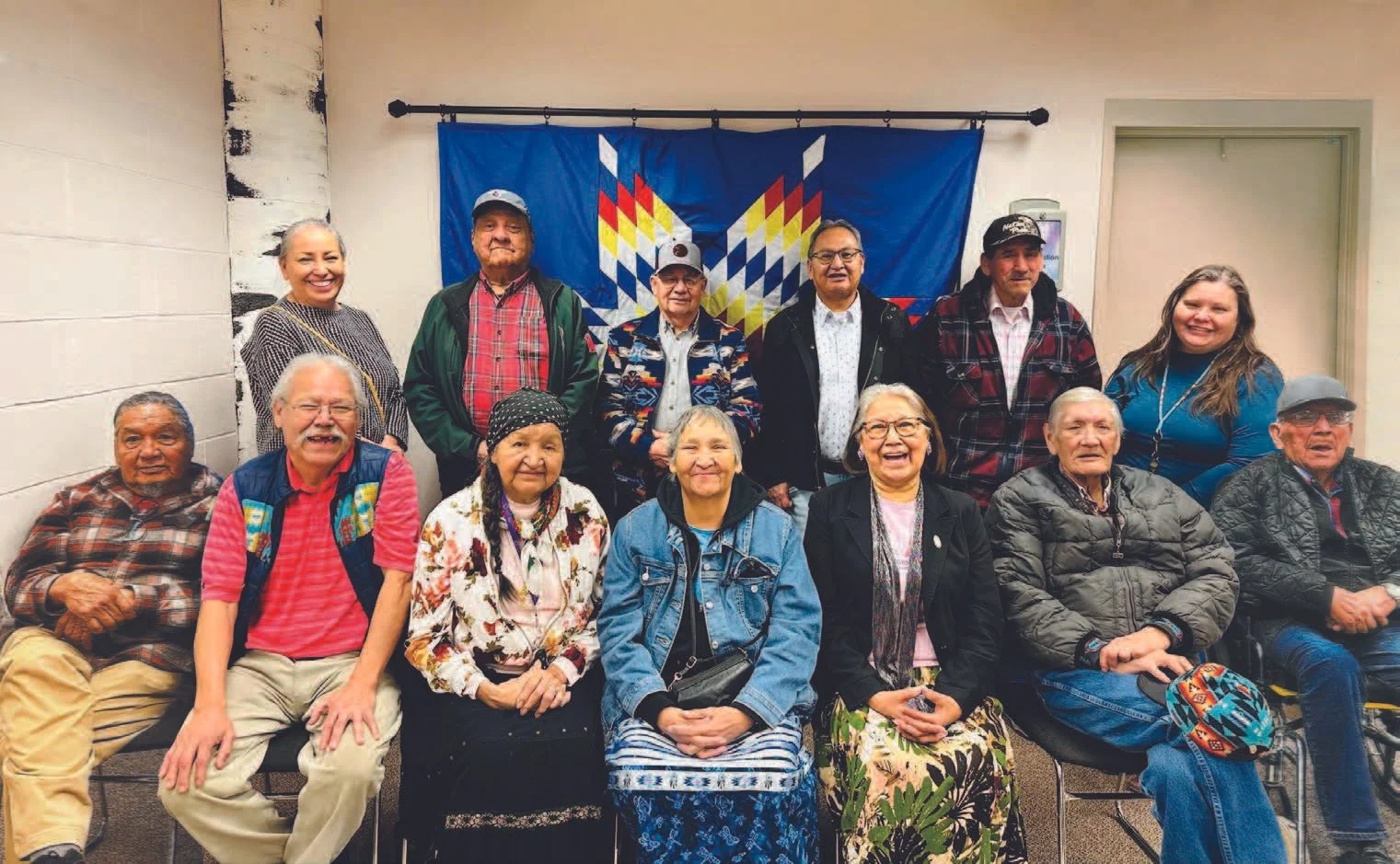 Group of Indigenous people and allies posing in front of a colorful Thunderbird flag at a cultural gathering.
