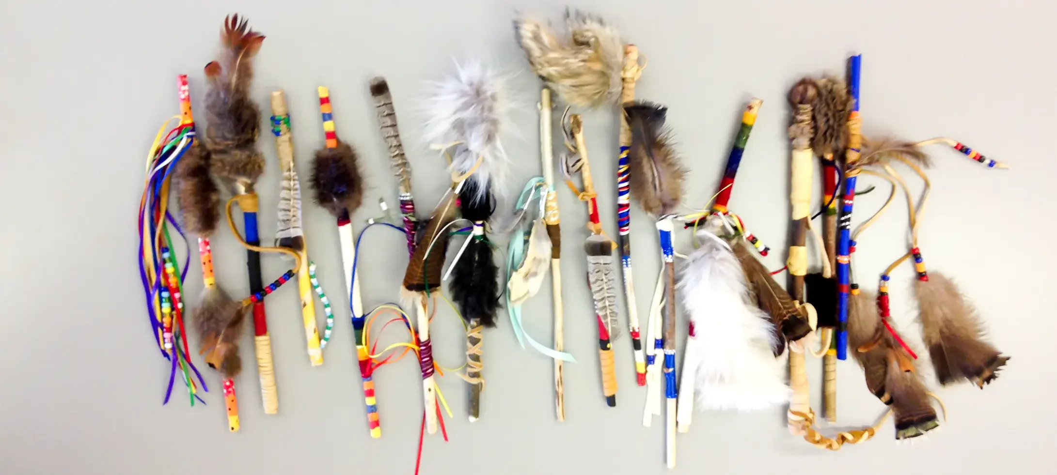A variety of handcrafted bulls, staffs and sticks decorated with colorful ribbons, feathers, beads, and other materials, arranged against a plain wall.
