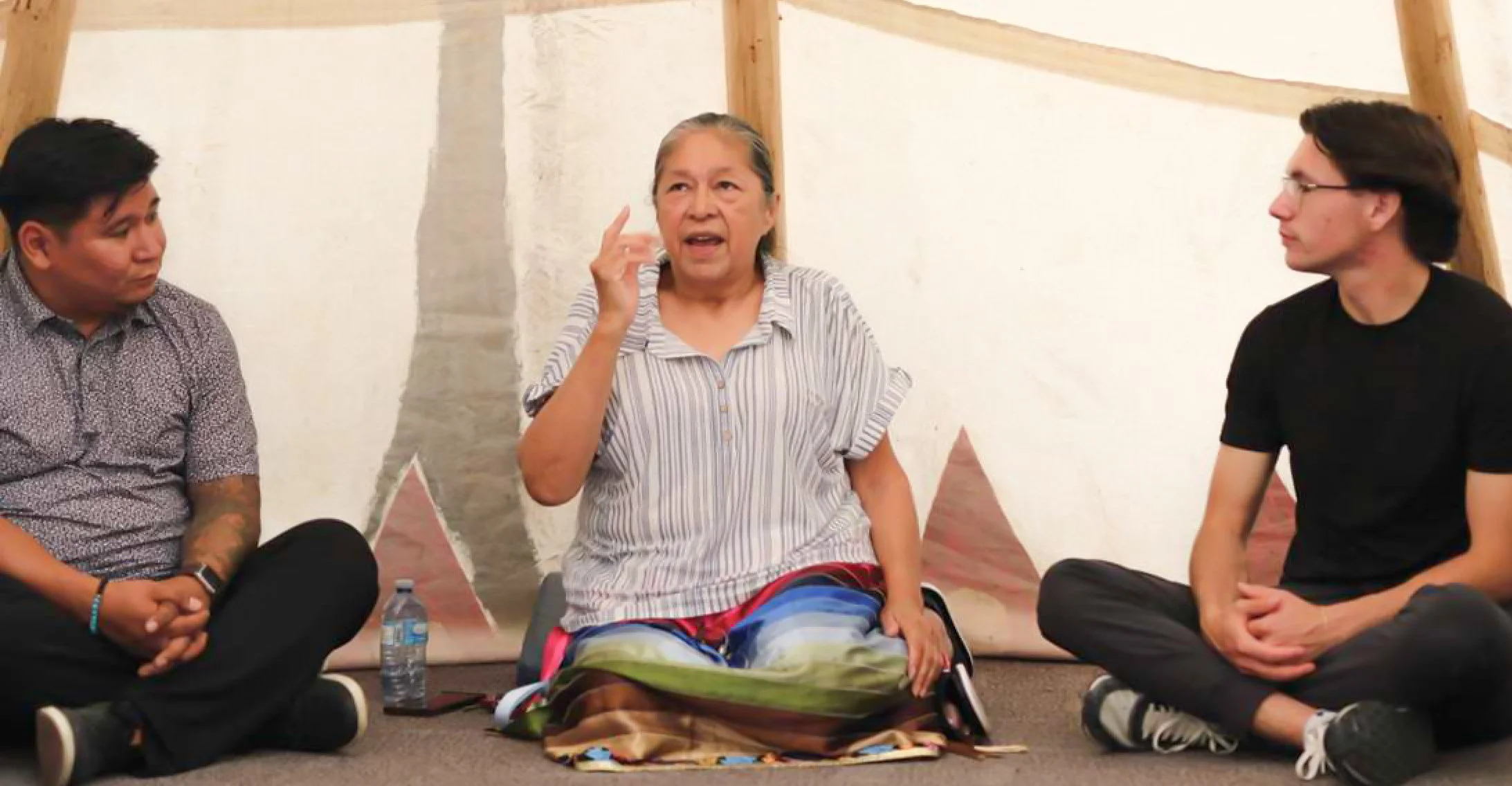 Three people sitting on the floor and having a discussion inside a tipi or teepee tent; an older woman in the middle is speaking, with two younger men on either side listening.