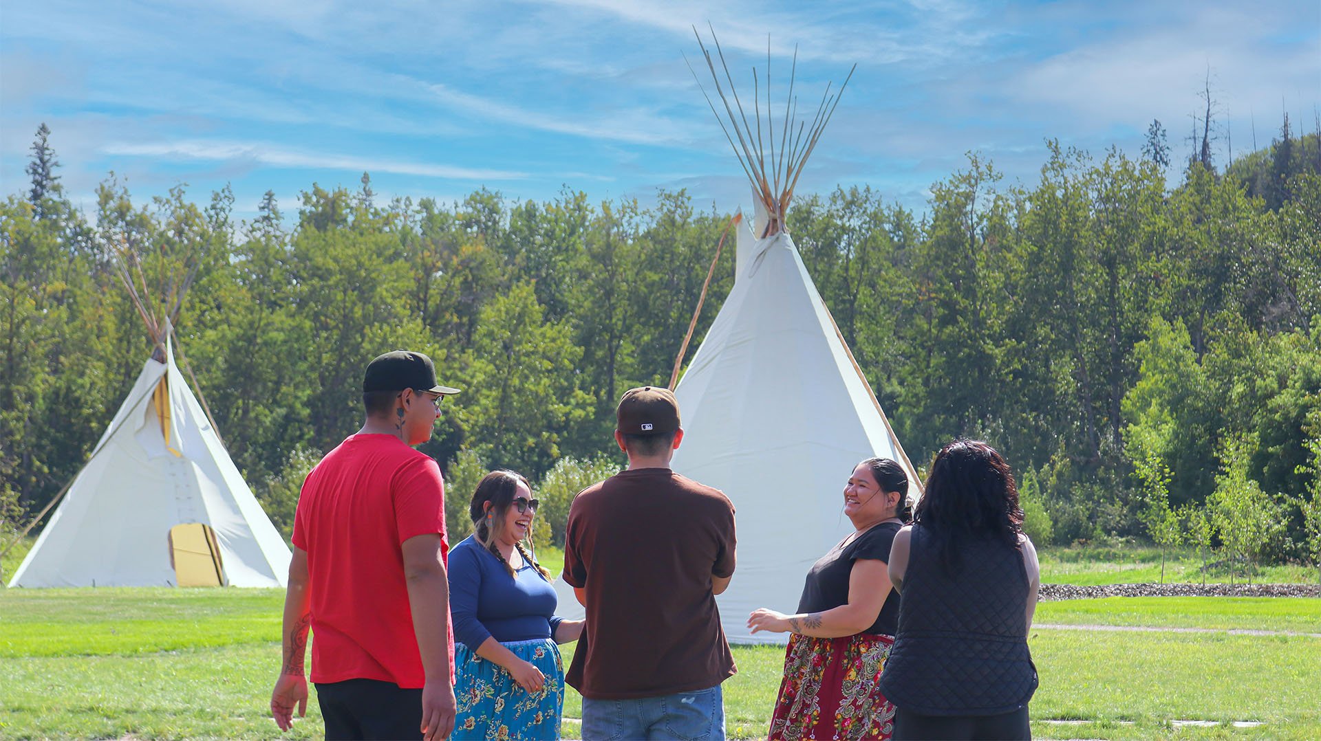 Group of five people standing on grass in front of two white tipi tents, engaging in conversation and smiling outdoors on a sunny day with green trees in the background.