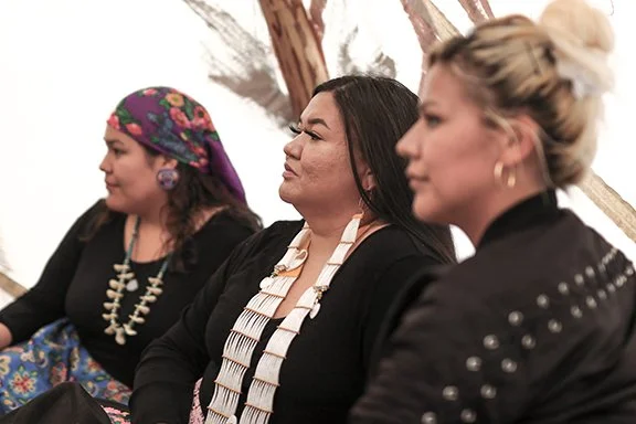 Three women sitting side by side, attentive, with a background of a tree.