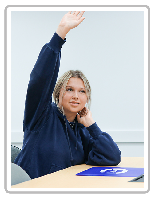 A young woman with blonde hair wearing a dark blue hoodie raising her hand in a classroom.