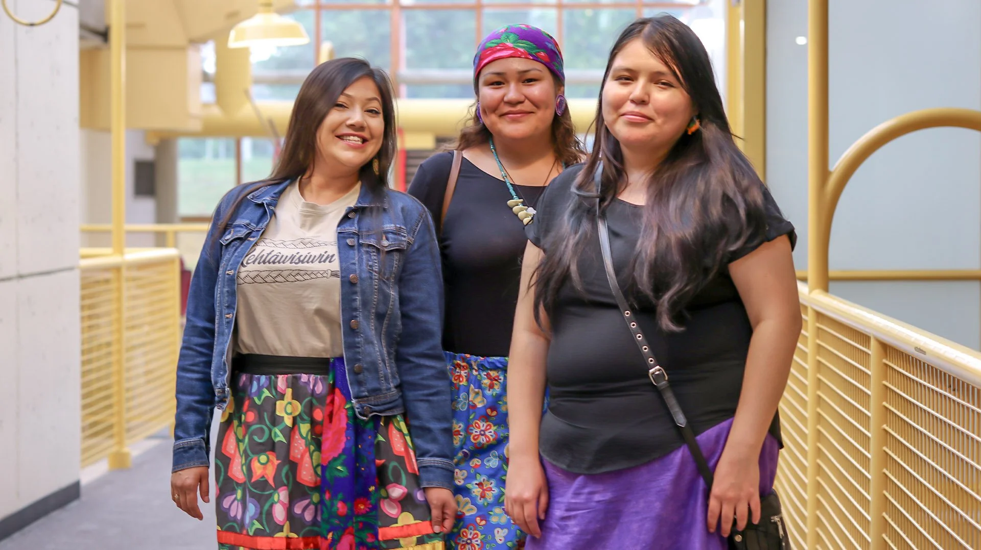 Three women standing together inside a building, smiling at the camera. The woman on the left wears a denim jacket over a graphic t-shirt and a colorful skirt. The woman in the middle wears a black top, a decorative necklace, a colorful headscarf, and a patterned skirt. The woman on the right wears a black t-shirt, a purple skirt, and has a black purse across her body.