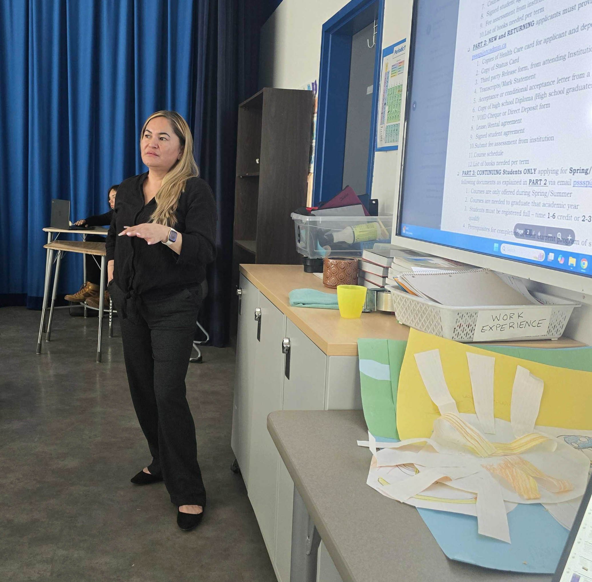 Woman standing with her hands gesturing, in a classroom or office setting, with another person sitting at a desk in the background, a large screen displaying text, and various papers and office supplies on the desks.