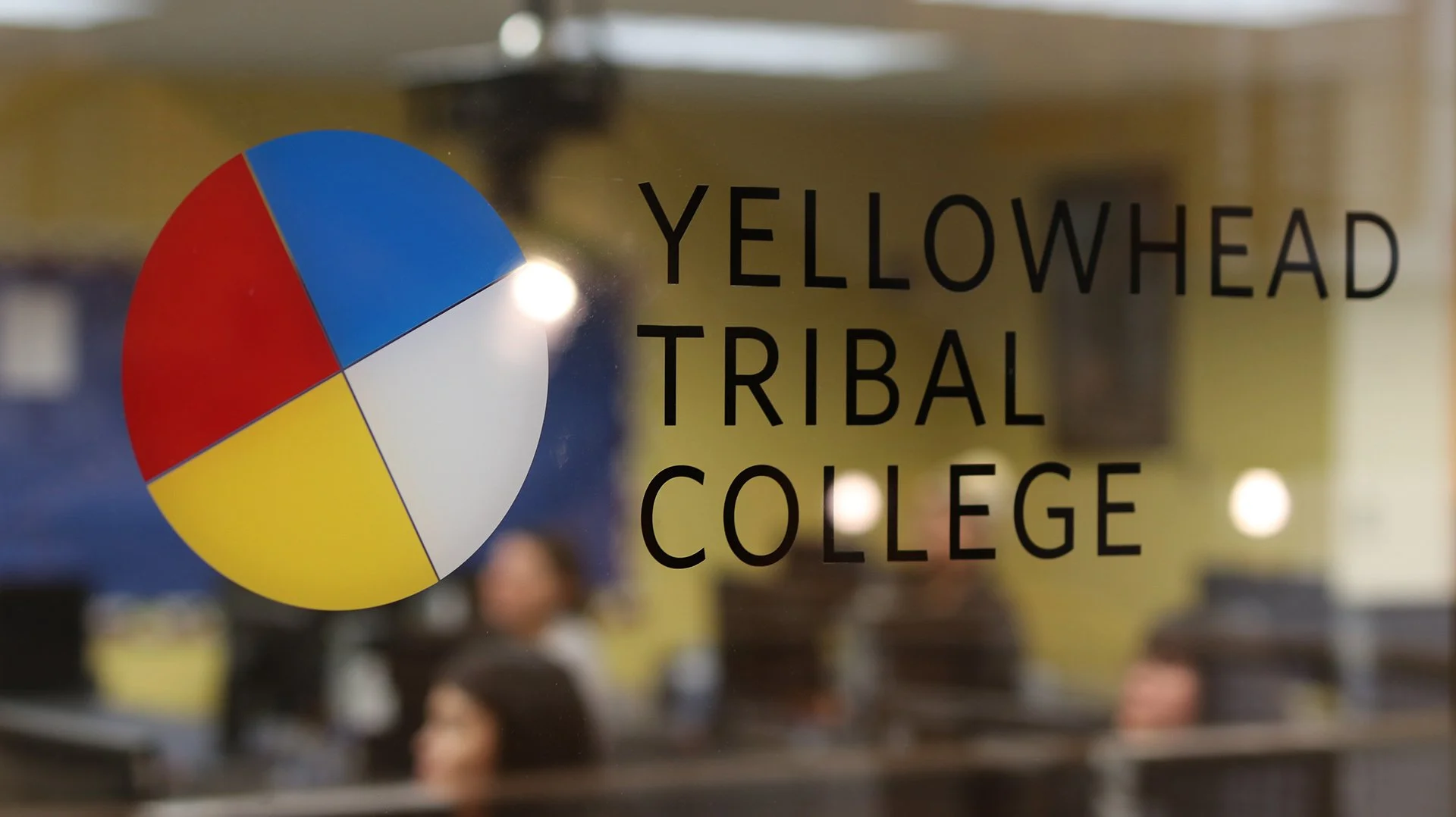 Glass door with the logo of Yellowhead Tribal College, featuring a colorful circular symbol and the college's name written in black letters. Inside, people are sitting and working at desks in a classroom or office setting.