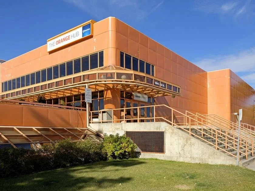 The Orange Hub, a modern orange-colored building with large windows and an exterior staircase, set against a blue sky.
