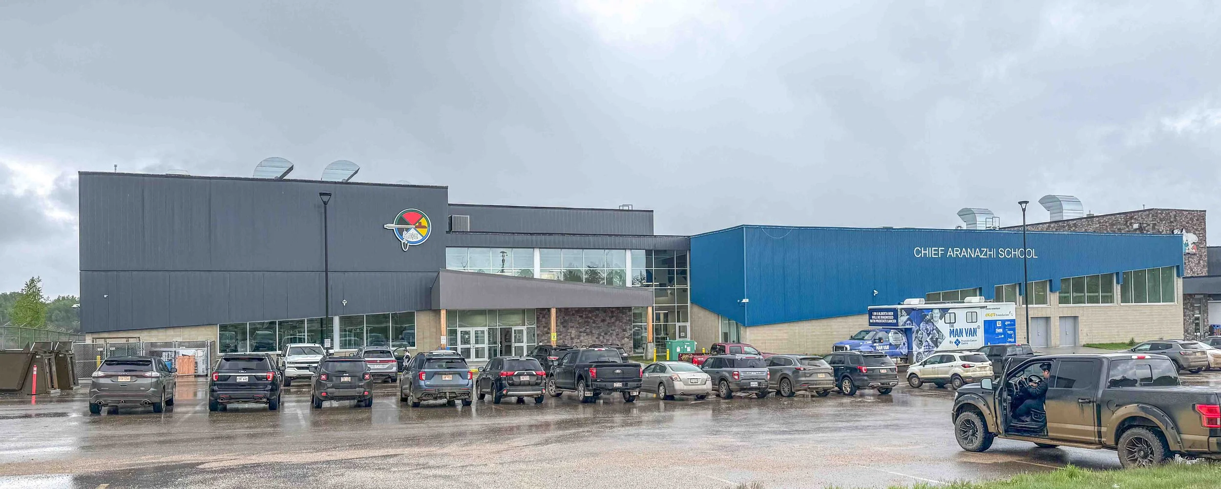 Front view of Chief Aranazhi School with a parking lot full of cars on a rainy day, overcast sky, and a person in a brown truck.