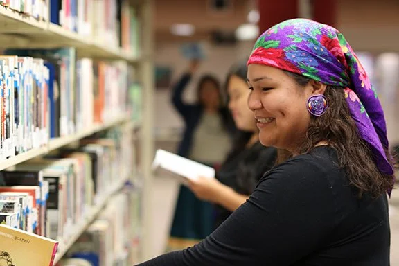 A woman with a colorful headscarf and purple earrings reading a book in a library, with other people browsing in the background.