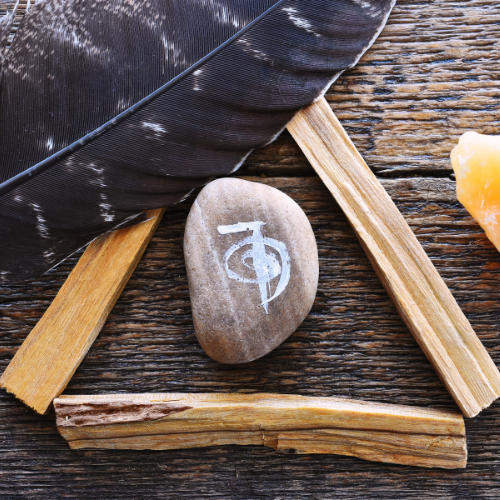 A stone with a white symbol, a black feather, and small wooden sticks on a rustic wooden surface.