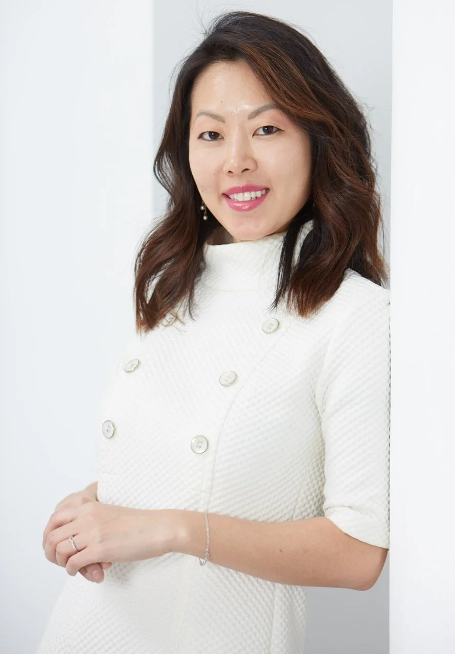 A woman with wavy brown hair, wearing a white textured dress with buttons, smiling and standing against a white background.