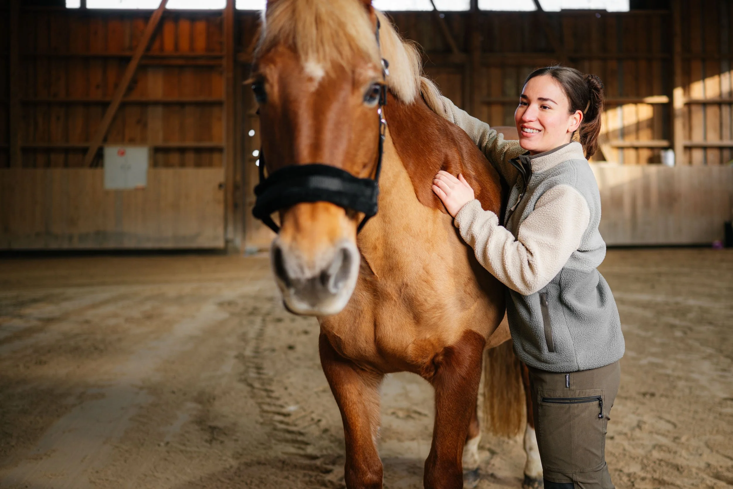 Junge Frau, die ein braunes Pferd in einer Stallhalle streichelt.