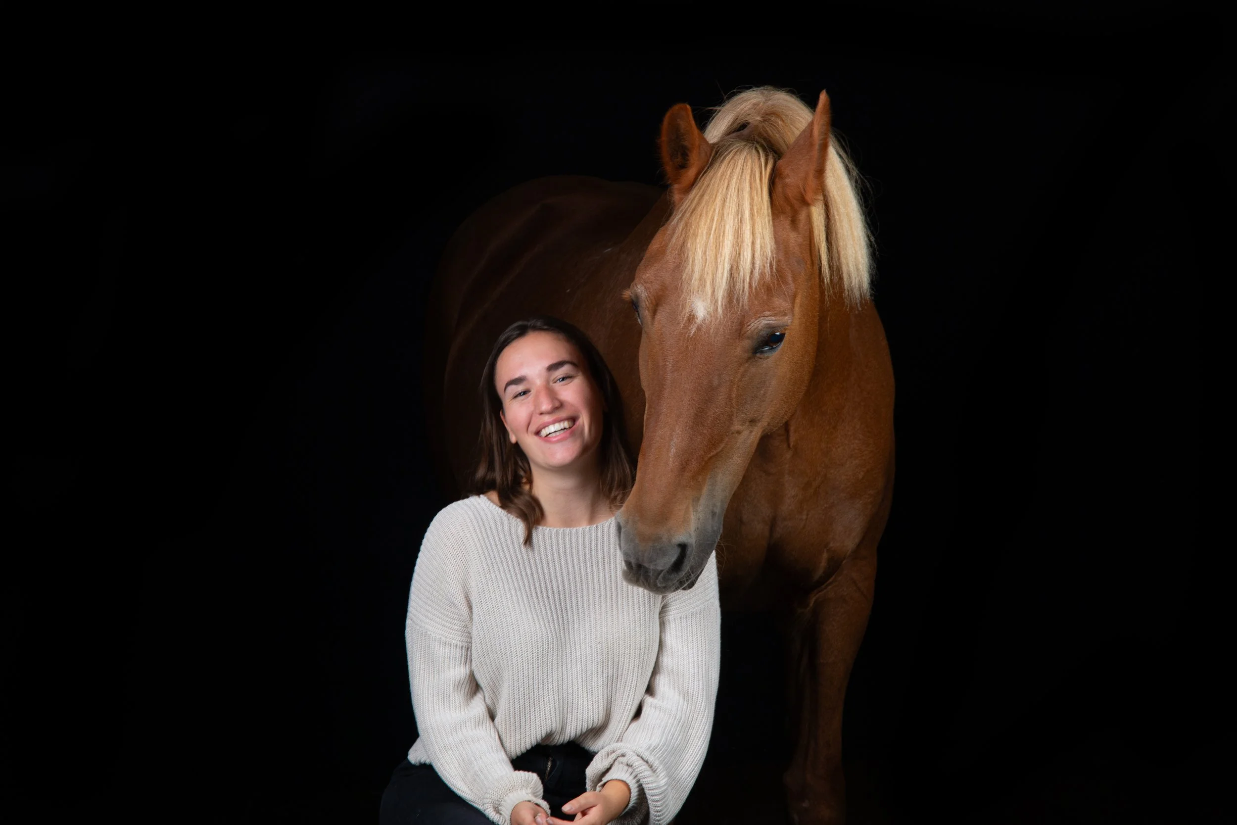 Junge Frau mit braunen Haaren, in einem weißen Pullover, neben einem braunen Pferd mit blonder Mähne vor schwarzem Hintergrund.