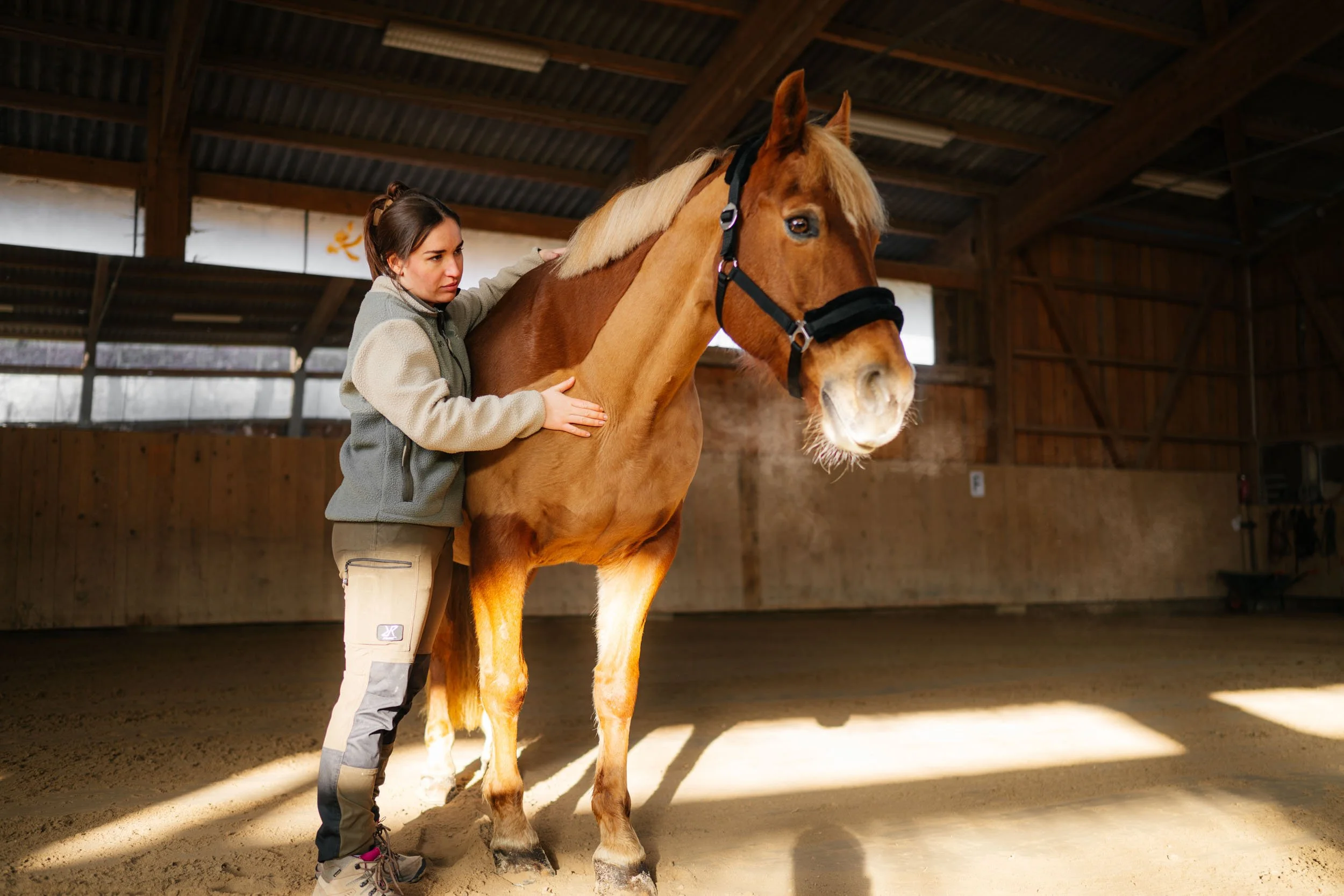 Junge Frau in Reithose und Jacke beim Abhören des Pferdes in einem Reithalle.